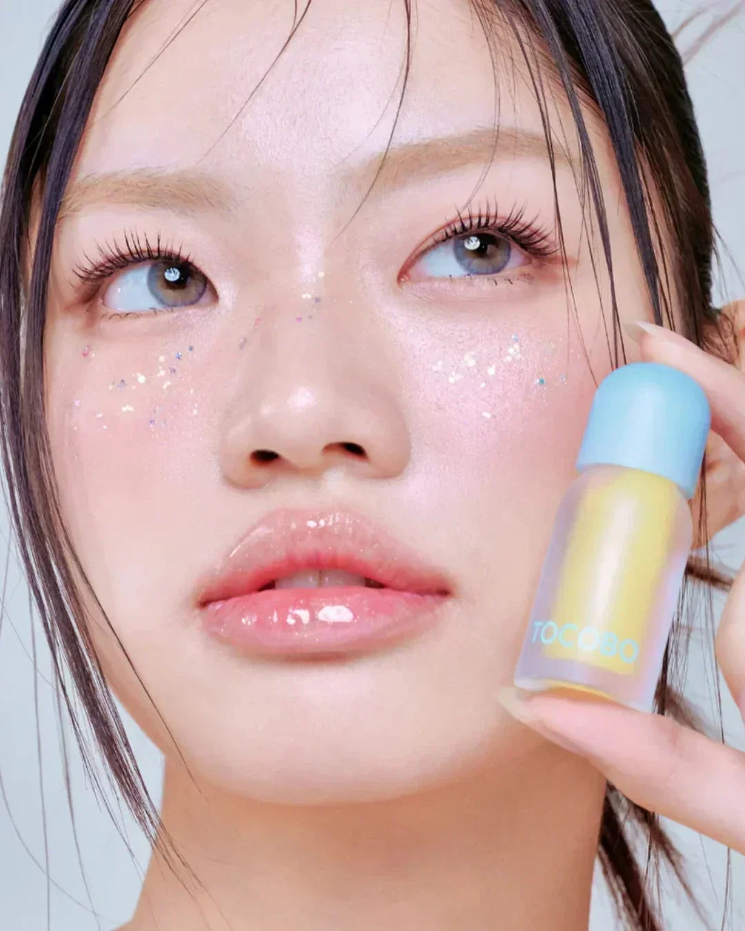 Close-up of young woman with glossy lips and glitter on cheeks holding Tocobo lip oil bottle
