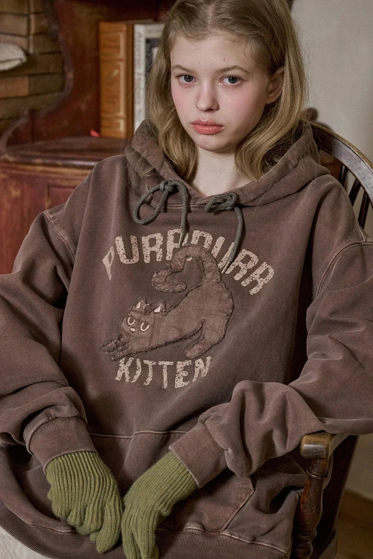 Young person in brown oversized hoodie with playful kitten graphic and text sitting on wooden chair