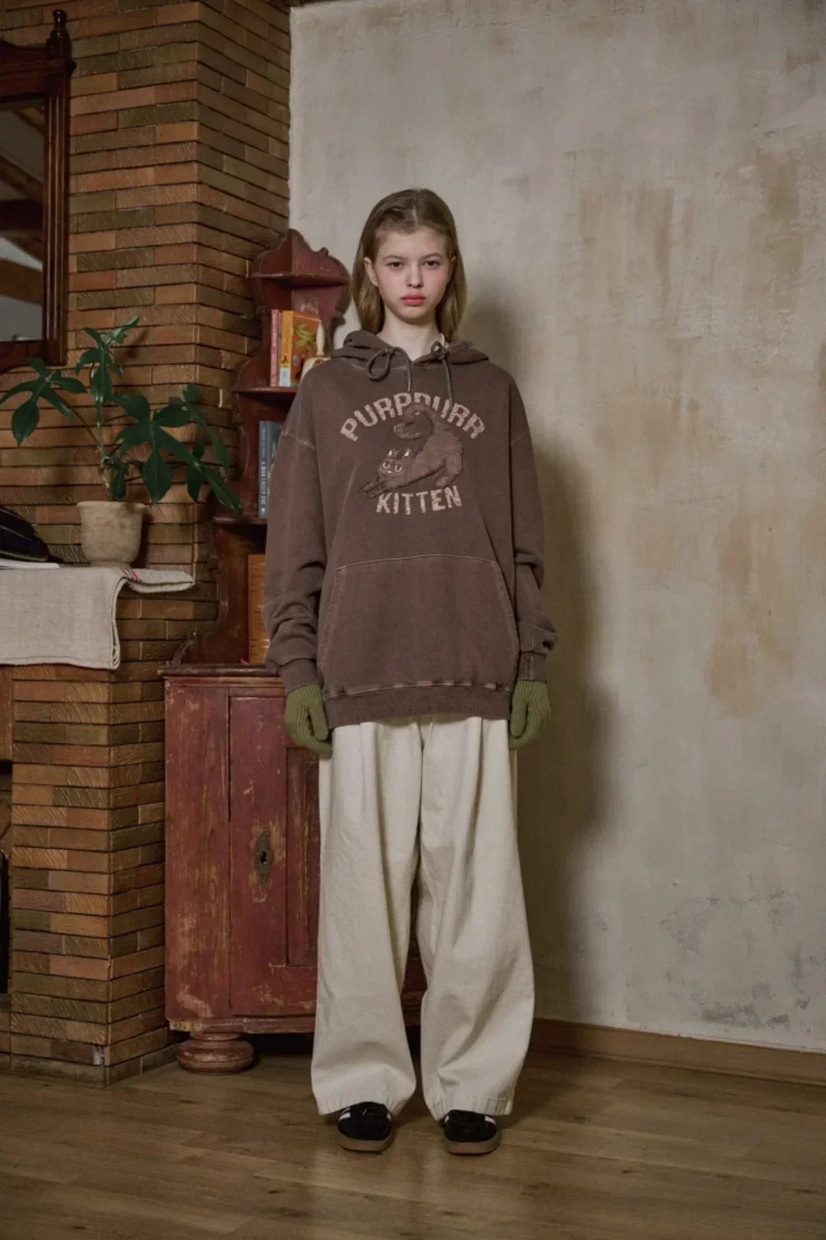 Young woman wearing brown kitten print hoodie, cream wide pants, green gloves, standing indoors