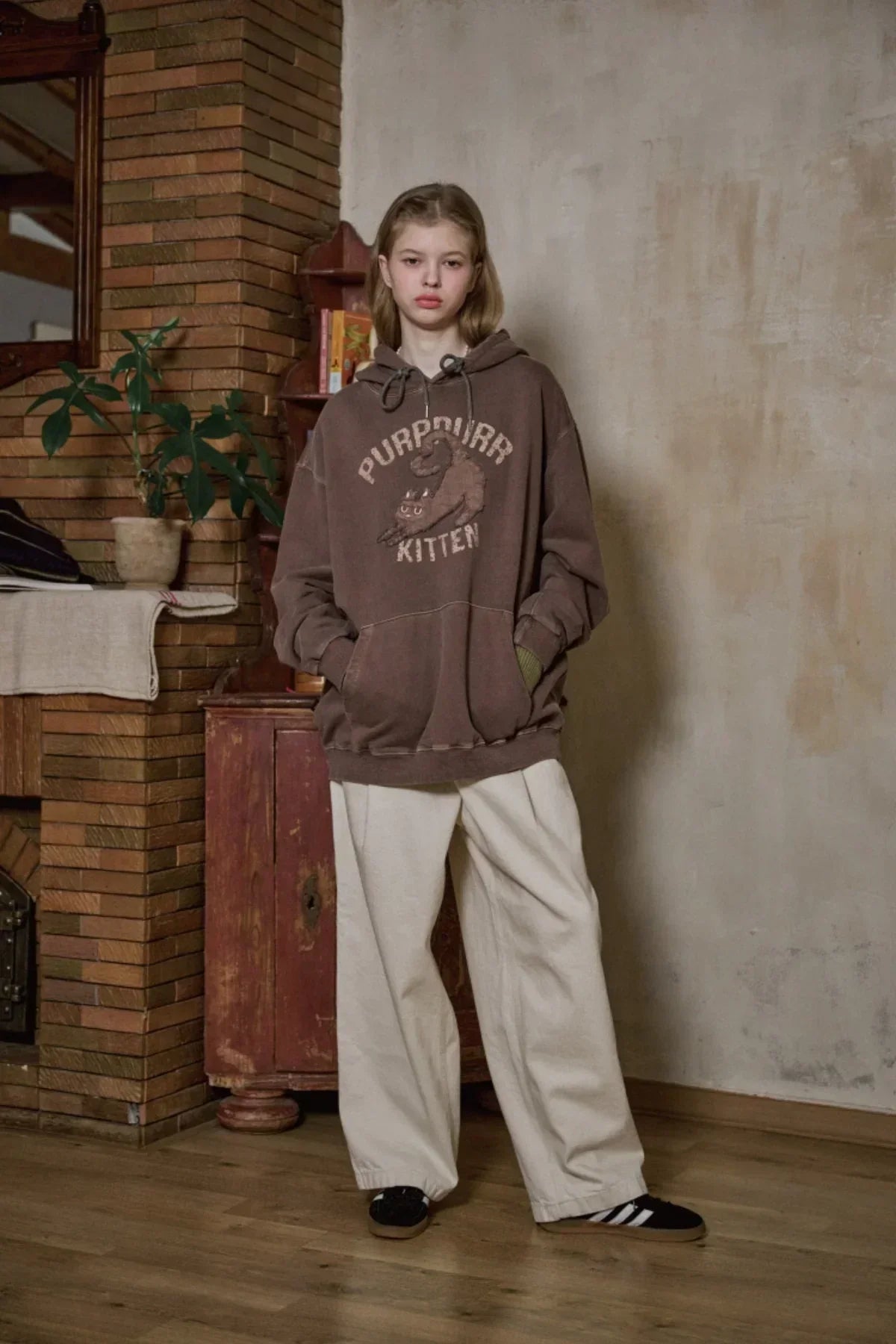 Young person wearing brown 'Purrrr Kitten' hoodie with graphic, white wide-leg pants indoors