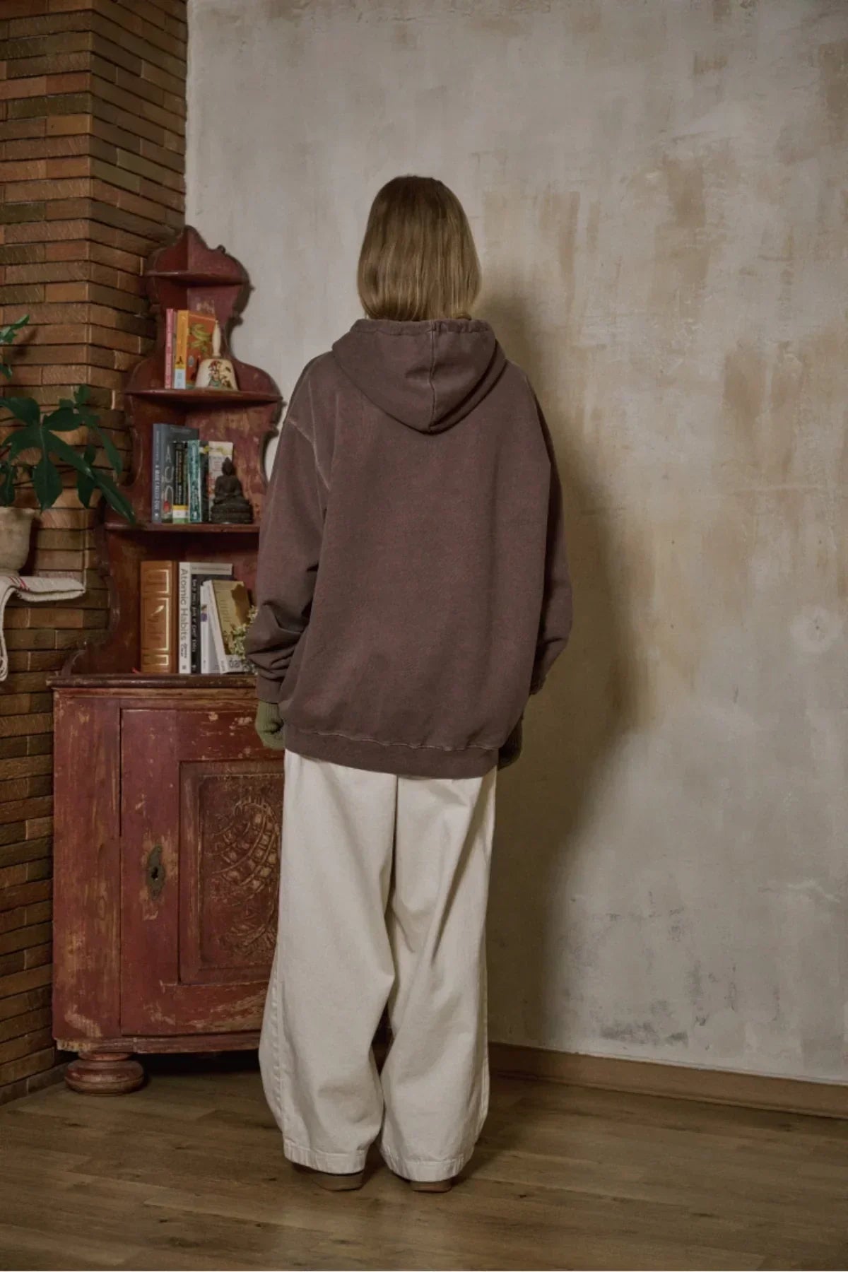 Person wearing brown hoodie and wide beige pants standing indoors by rustic wooden cabinet with books