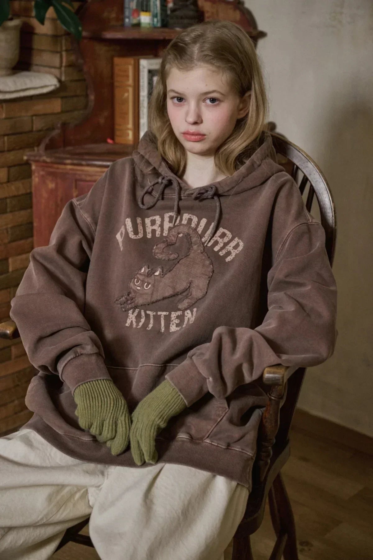 Young woman wearing brown kitten graphic hoodie and green knit gloves sitting in a wooden chair indoors