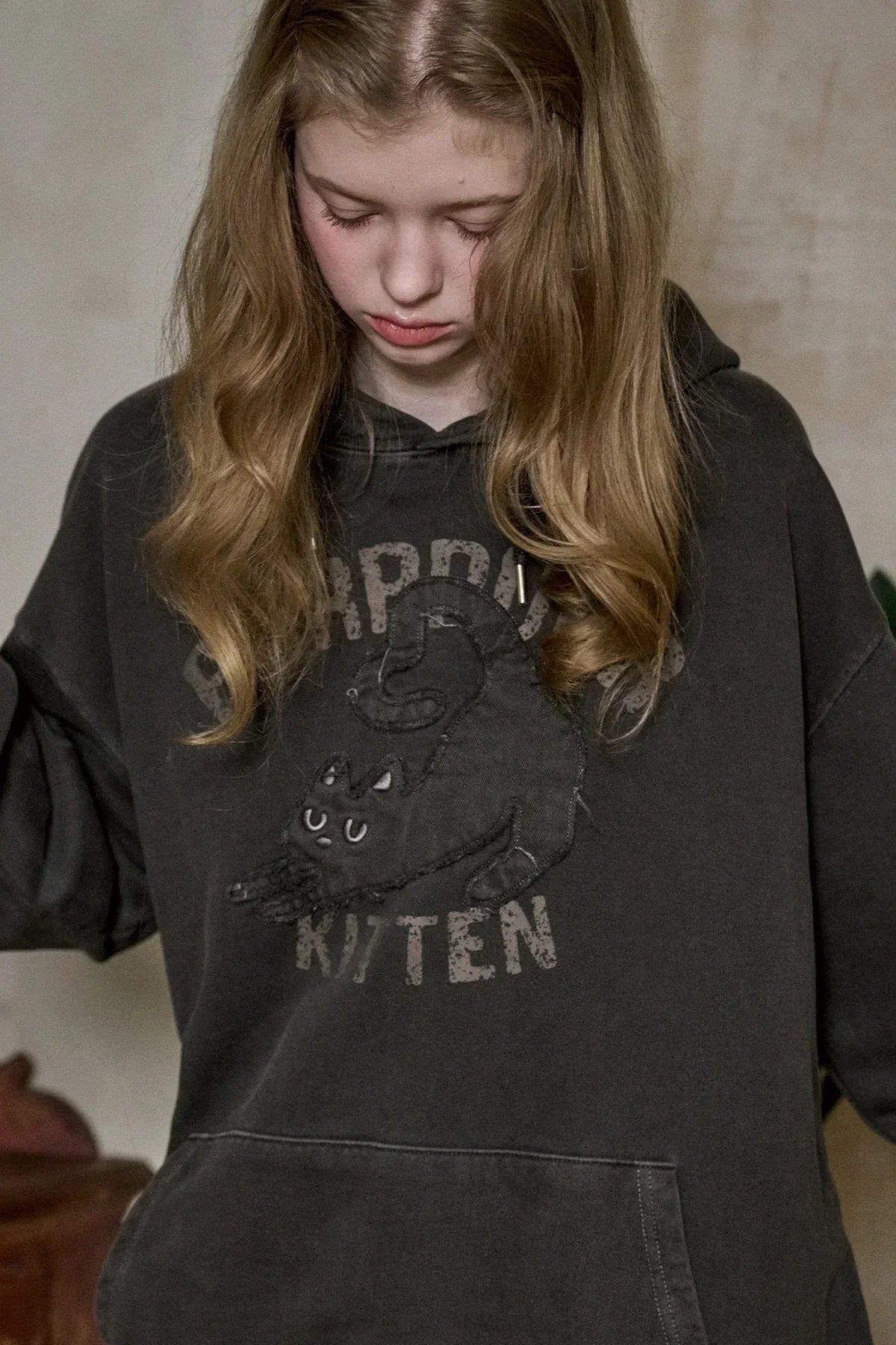 Young girl with long blonde hair wearing a black hoodie featuring a stretching black cat and text