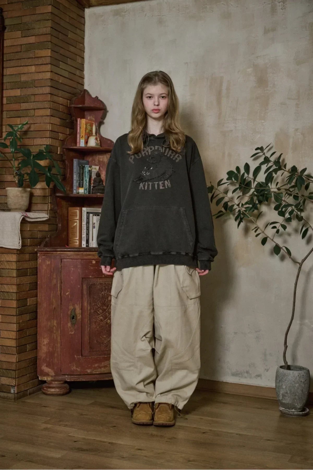 young woman wearing black kitten graphic hoodie and beige cargo pants standing indoors near vintage cabinet and potted plant