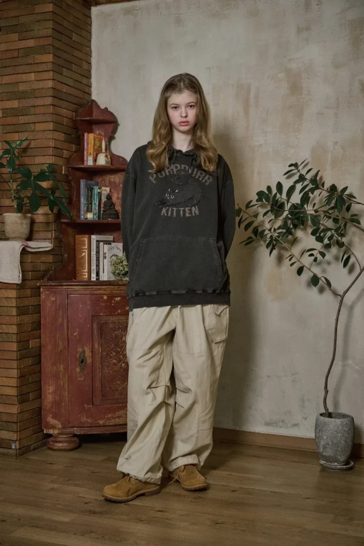 Young woman wearing black kitten graphic hoodie, beige cargo pants, and brown boots indoors