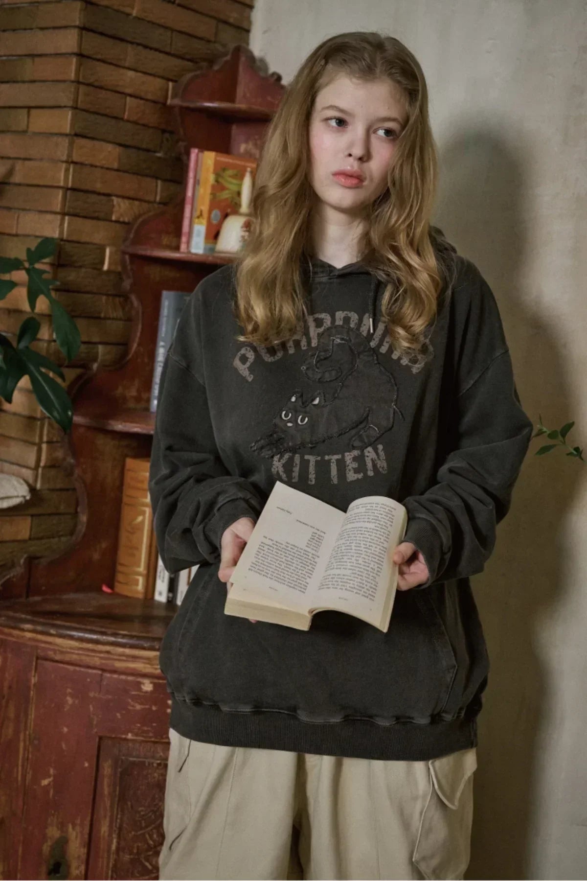 Young woman in black kitten graphic hoodie reading a book indoors by rustic bookshelf