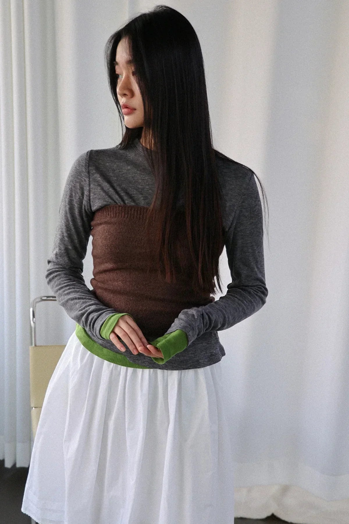 Woman with long dark hair wearing a gray long-sleeve top layered with a brown knit tube top and white skirt