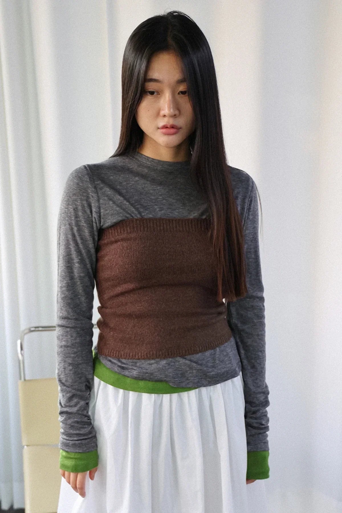 Woman wearing brown knit tube top over gray long sleeve shirt with green cuffs and white skirt indoors