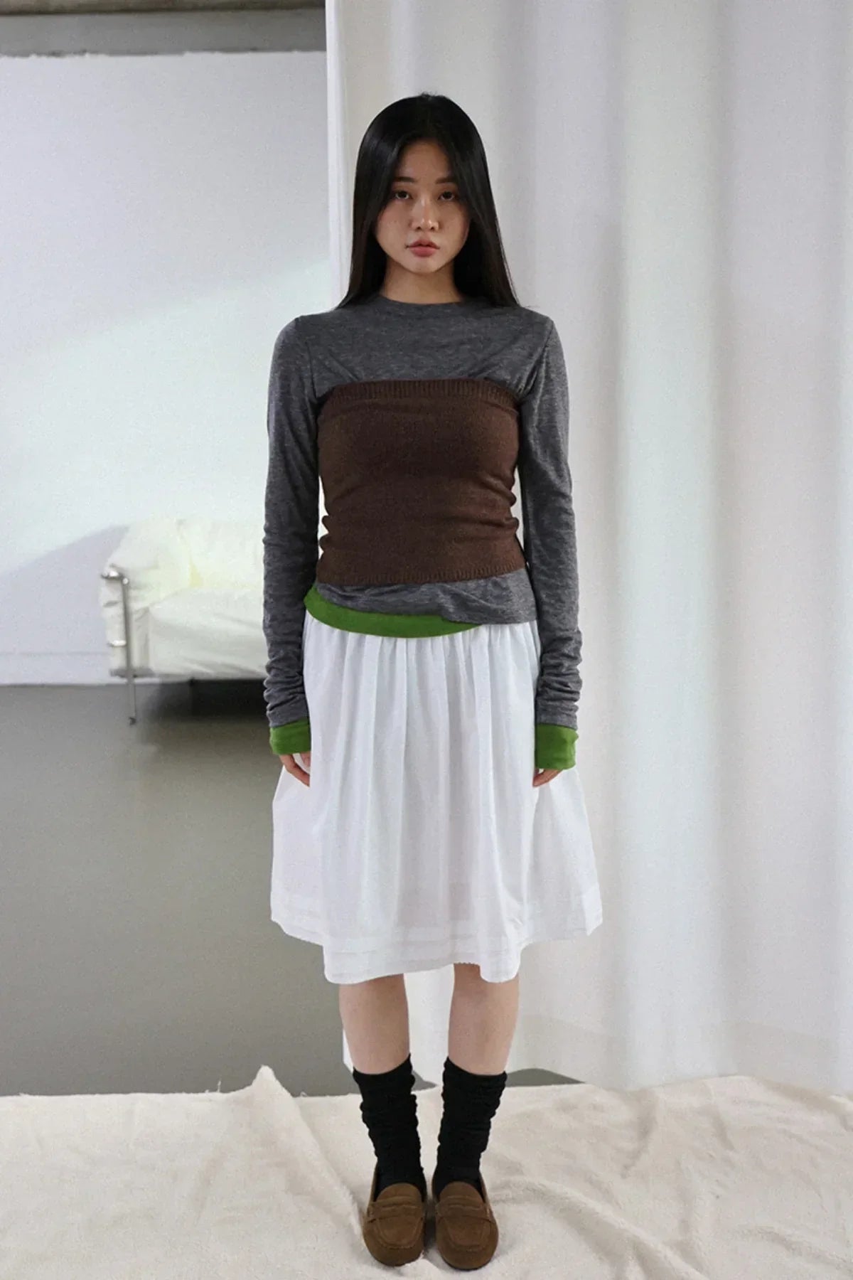 Woman wearing brown knit tube top over gray long sleeve with green cuffs, white skirt, black socks, brown loafers
