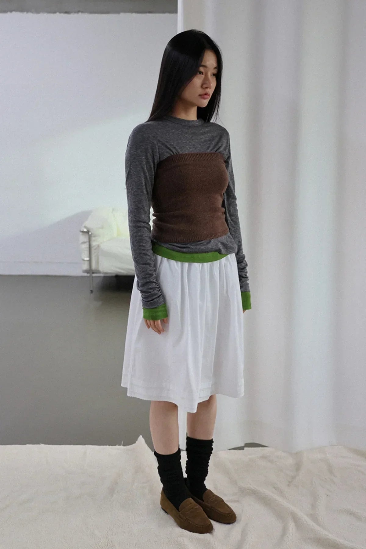 Model wearing brown knit tube top layered over gray long sleeve shirt with green cuffs, white skirt, and brown loafers