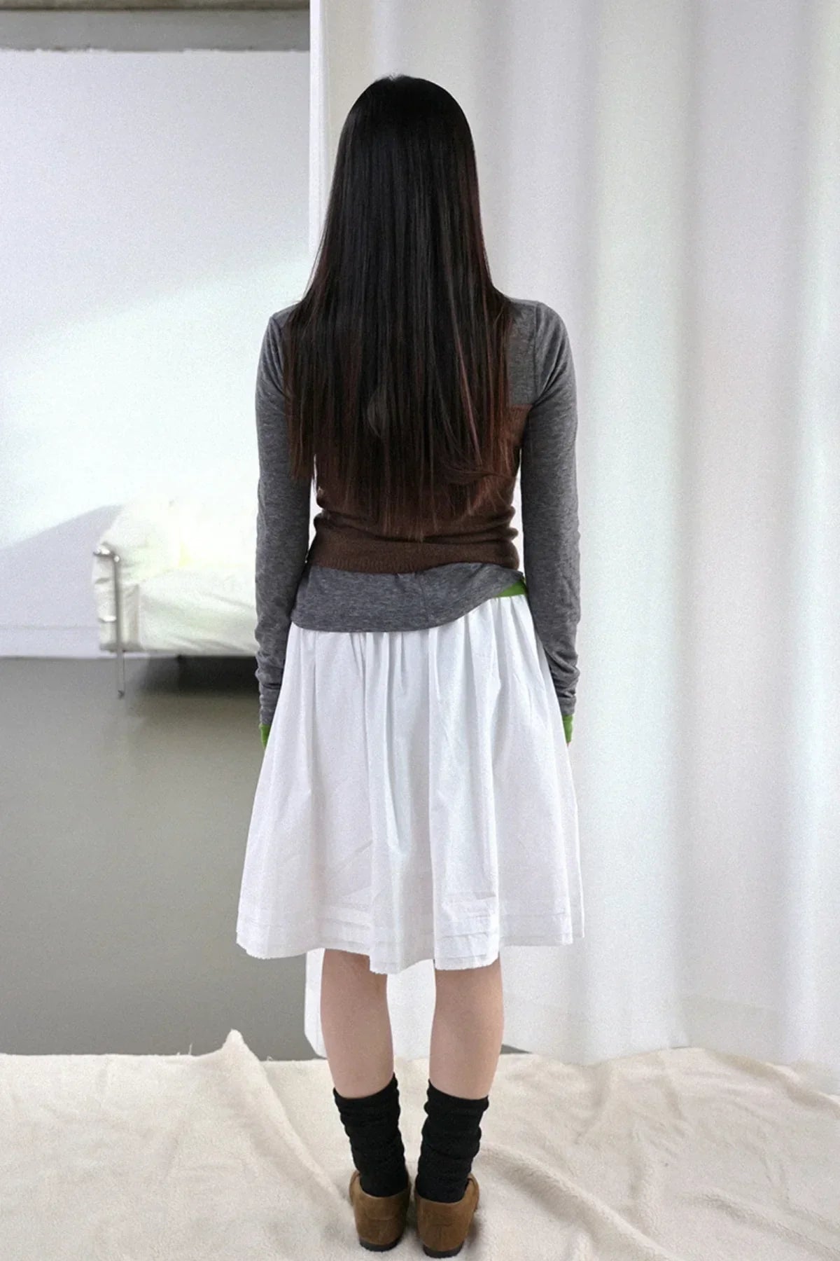 Woman with long hair wearing brown knit top over gray shirt and white skirt, standing indoors