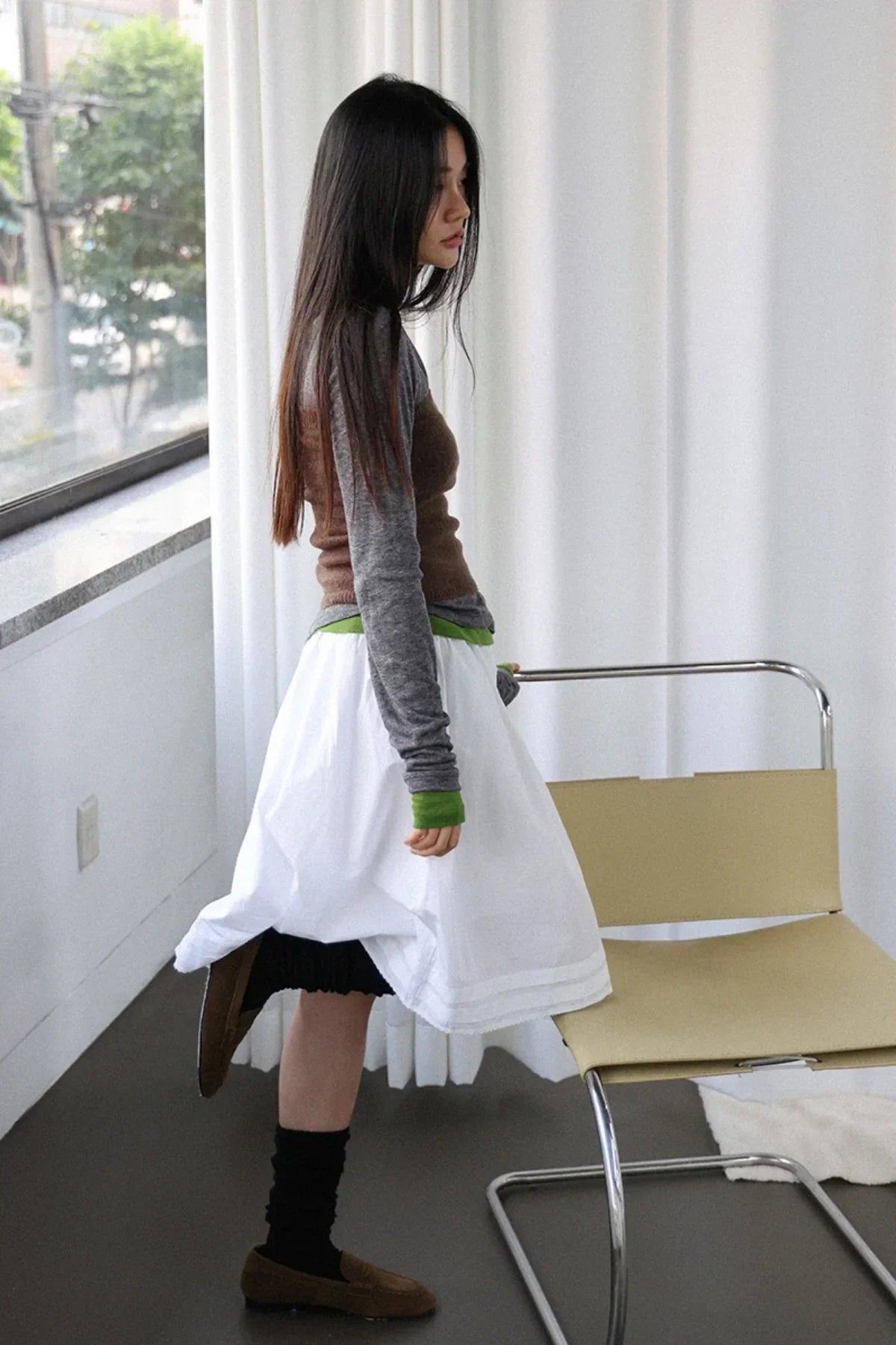 Woman in layered brown knit top, gray long sleeves, white skirt, and black socks with brown loafers indoors