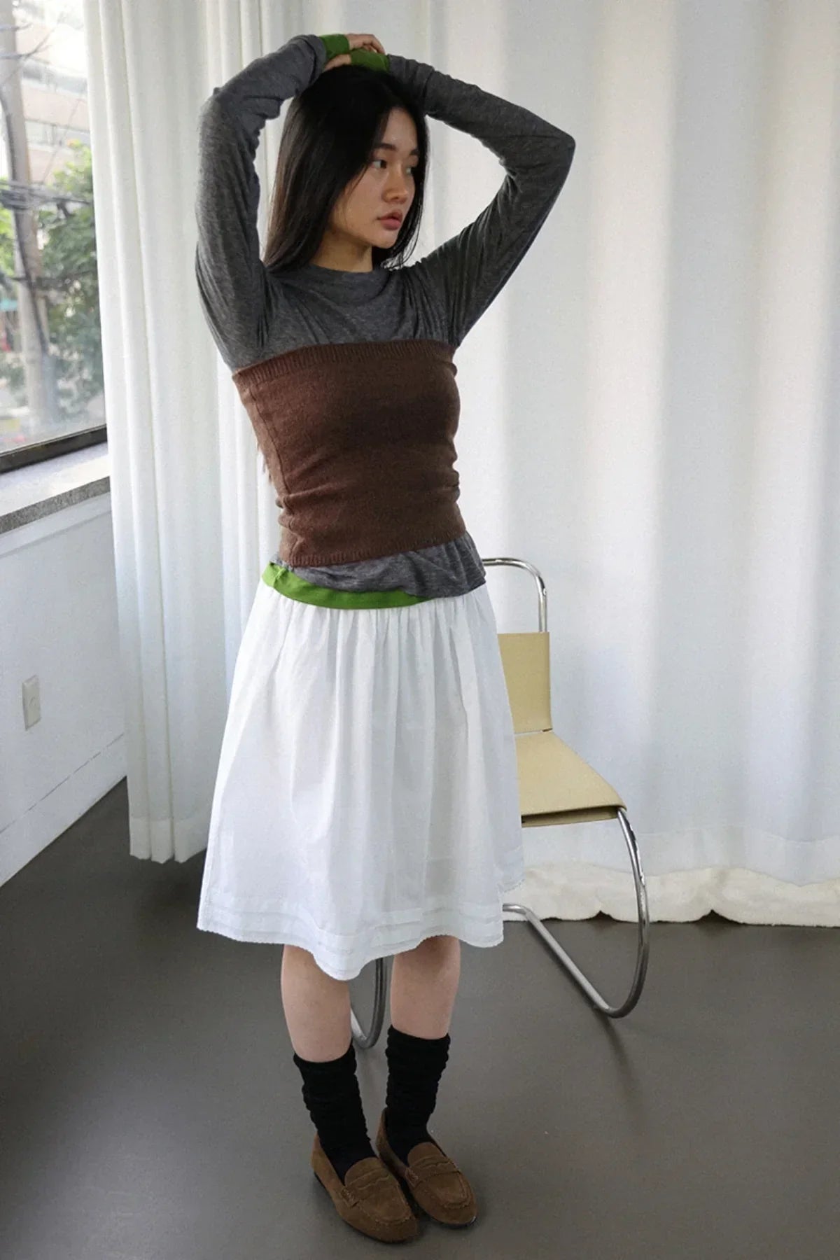 Young woman posing in brown knit tube top over gray long sleeve shirt with white skirt and black socks indoors