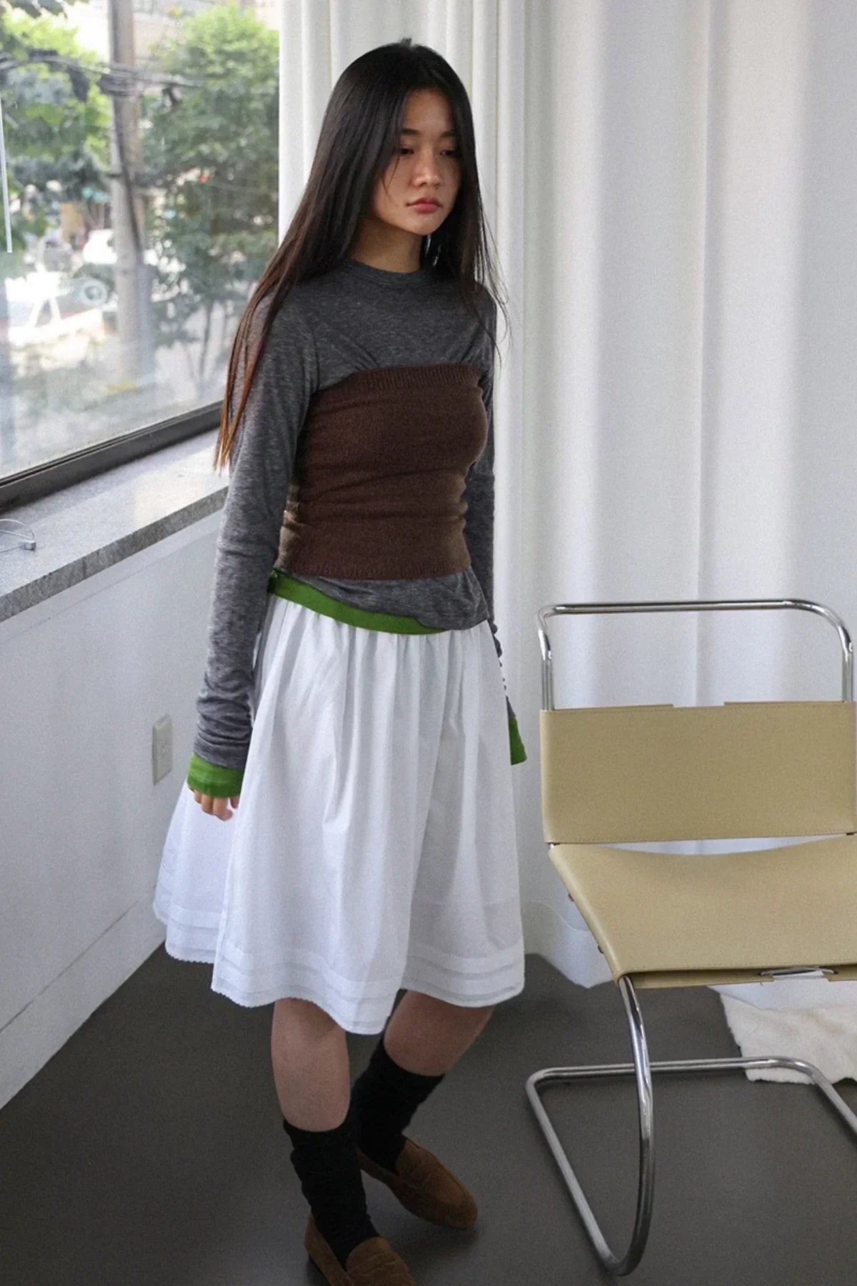 Woman wearing gray long-sleeve top with brown knit tube top, white skirt, and brown loafers in bright room