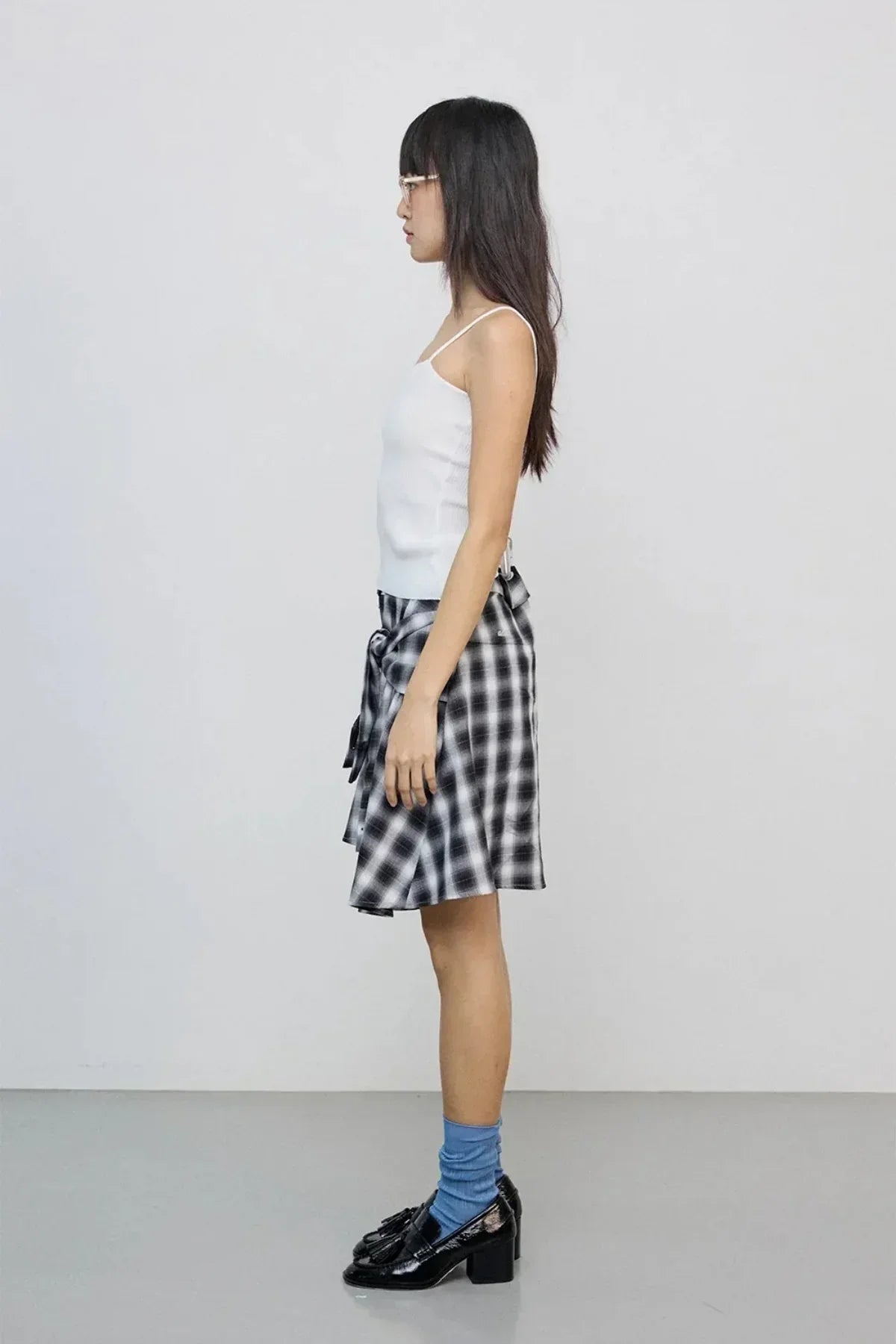 Side view of woman wearing white tank top, black and white checkered skirt, blue socks, and black loafers