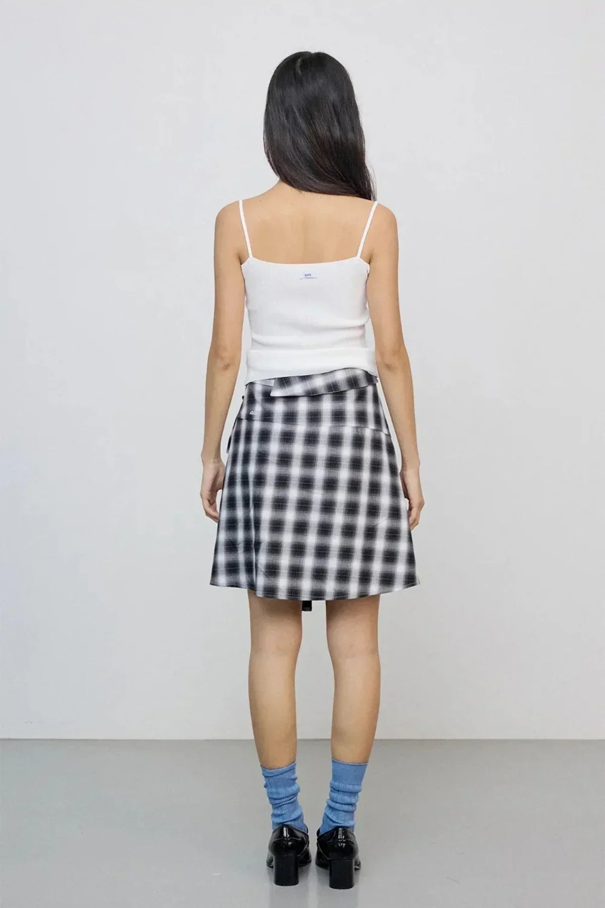 Woman wearing a black and white check skirt with a white sleeveless top, blue socks, and black shoes, standing indoors