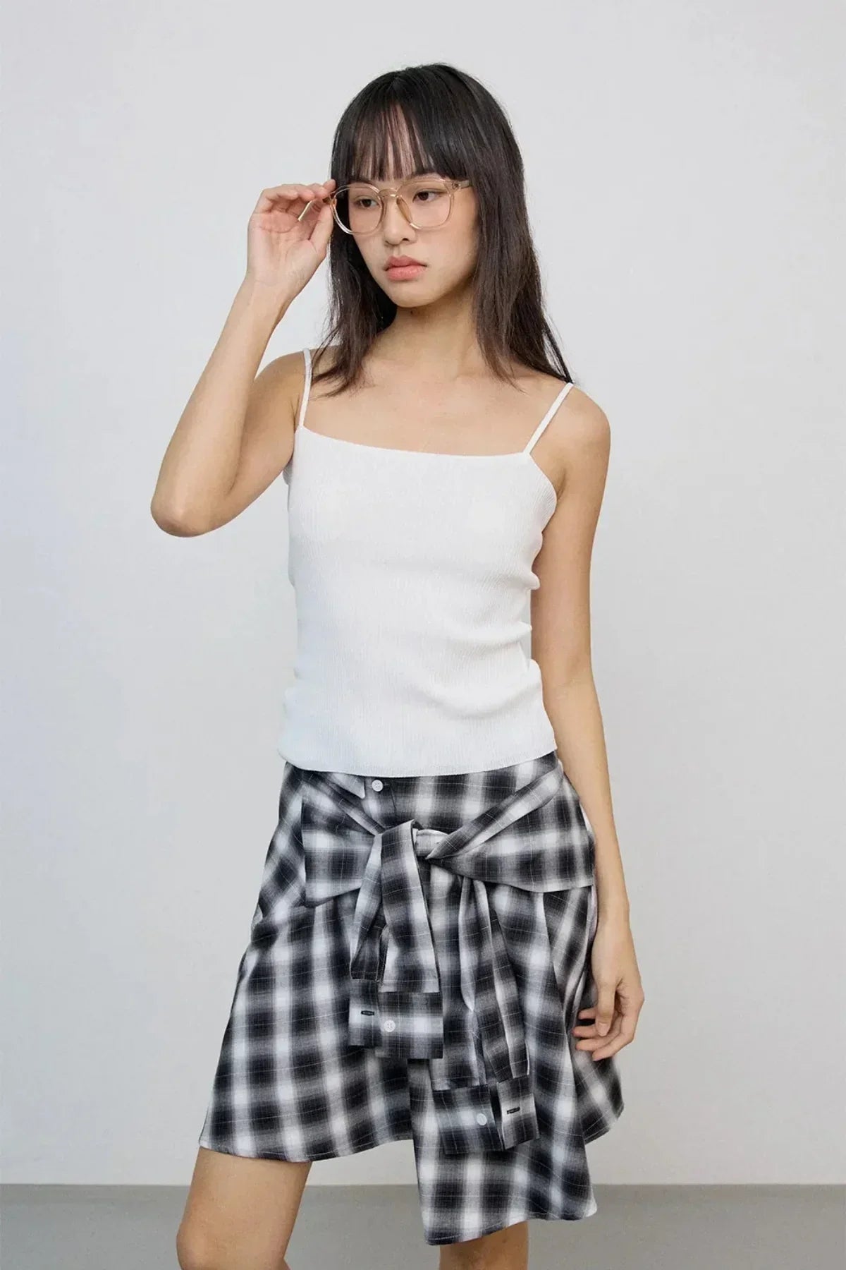 Young woman wearing clear glasses, white sleeveless top, and black white checkered skirt in studio