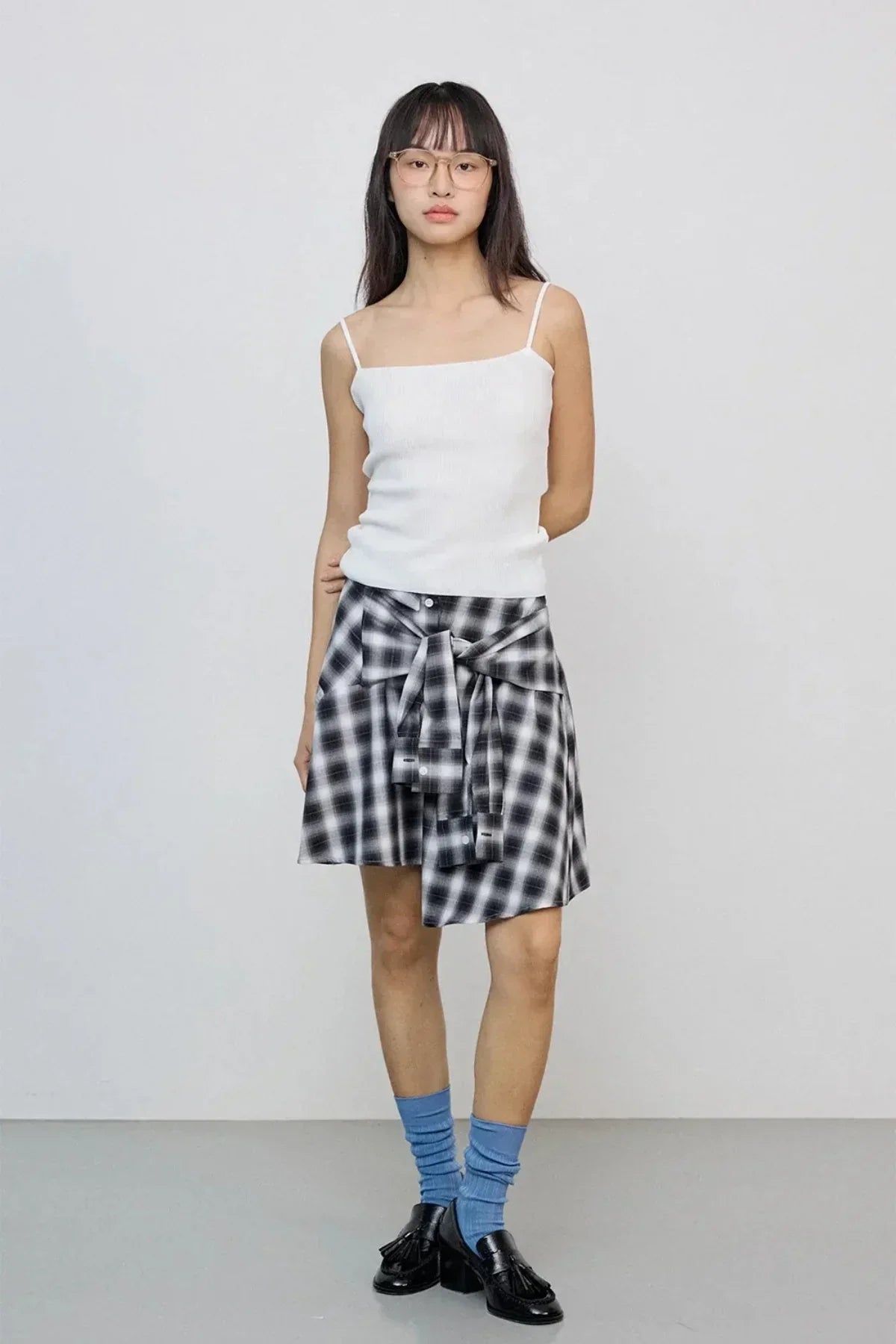 Young woman wearing a white spaghetti strap top, black and white checkered skirt, blue socks, and black loafers