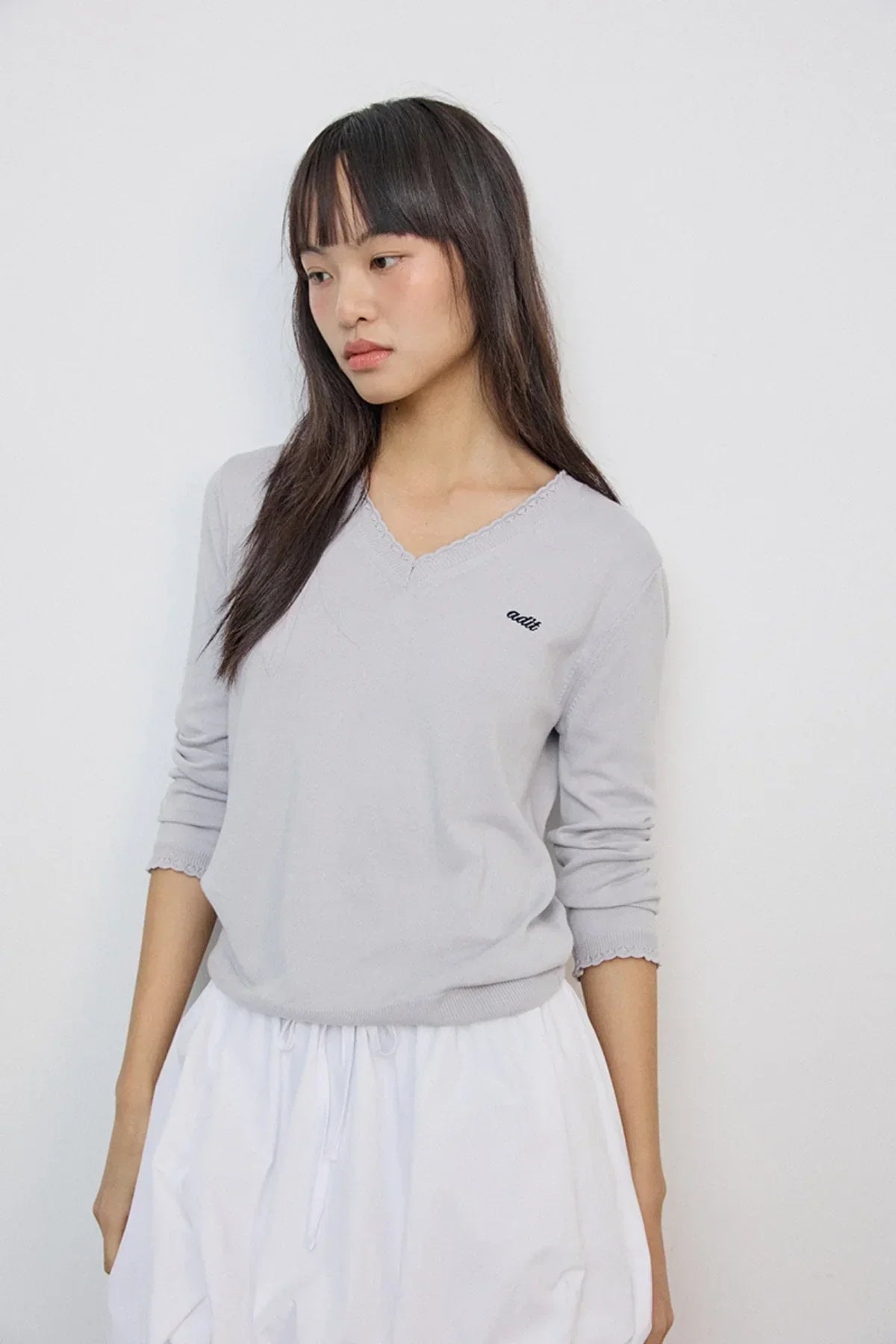 Asian woman wearing light gray V-neck knit sweater with scalloped edges and white skirt
