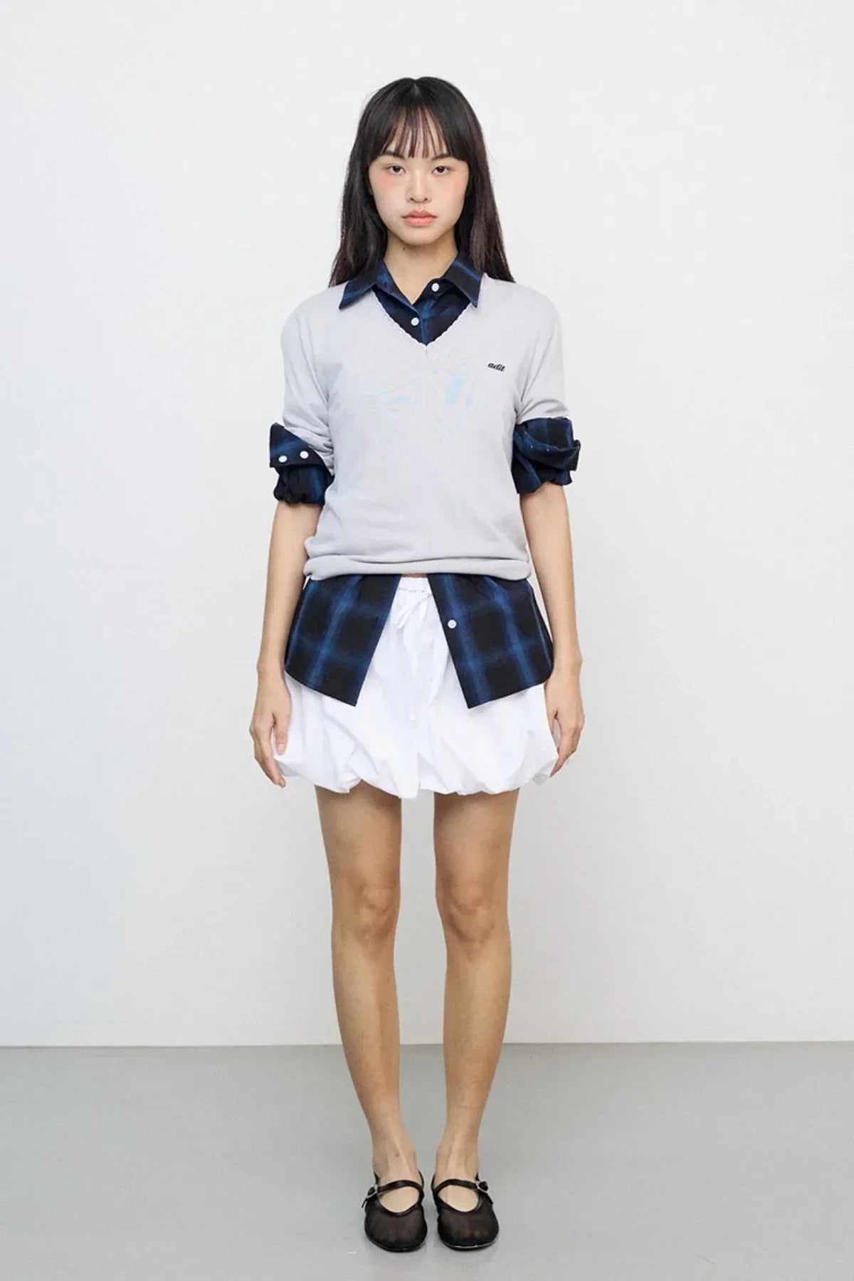 Asian woman in gray v-neck knit sweater layered over blue plaid shirt and white skirt with black flats