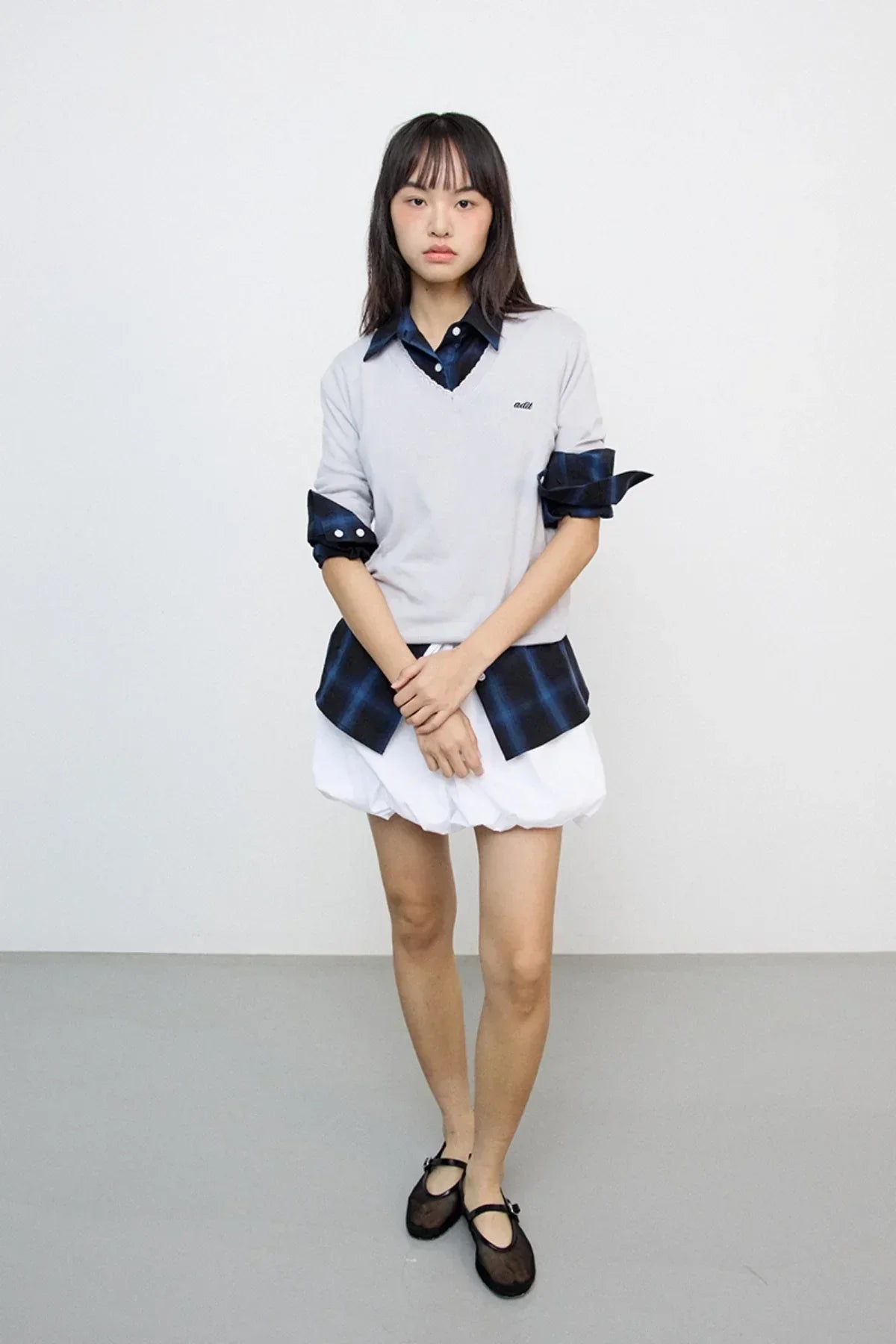 Asian female model wearing gray V-neck knit sweater over blue plaid shirt and white bubble skirt with black flats