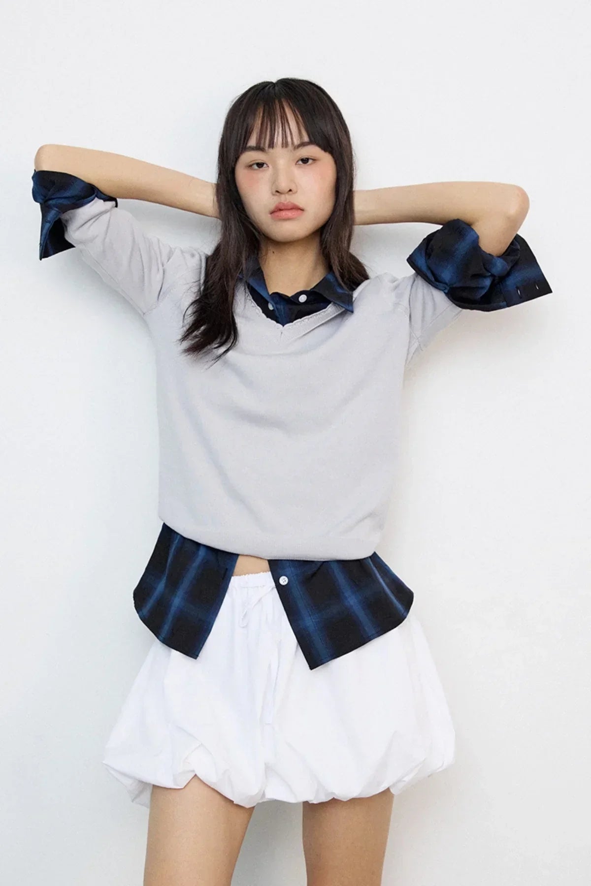 Young woman in layered outfit with gray lace v-neck knit sweater, blue plaid shirt, and white bubble skirt