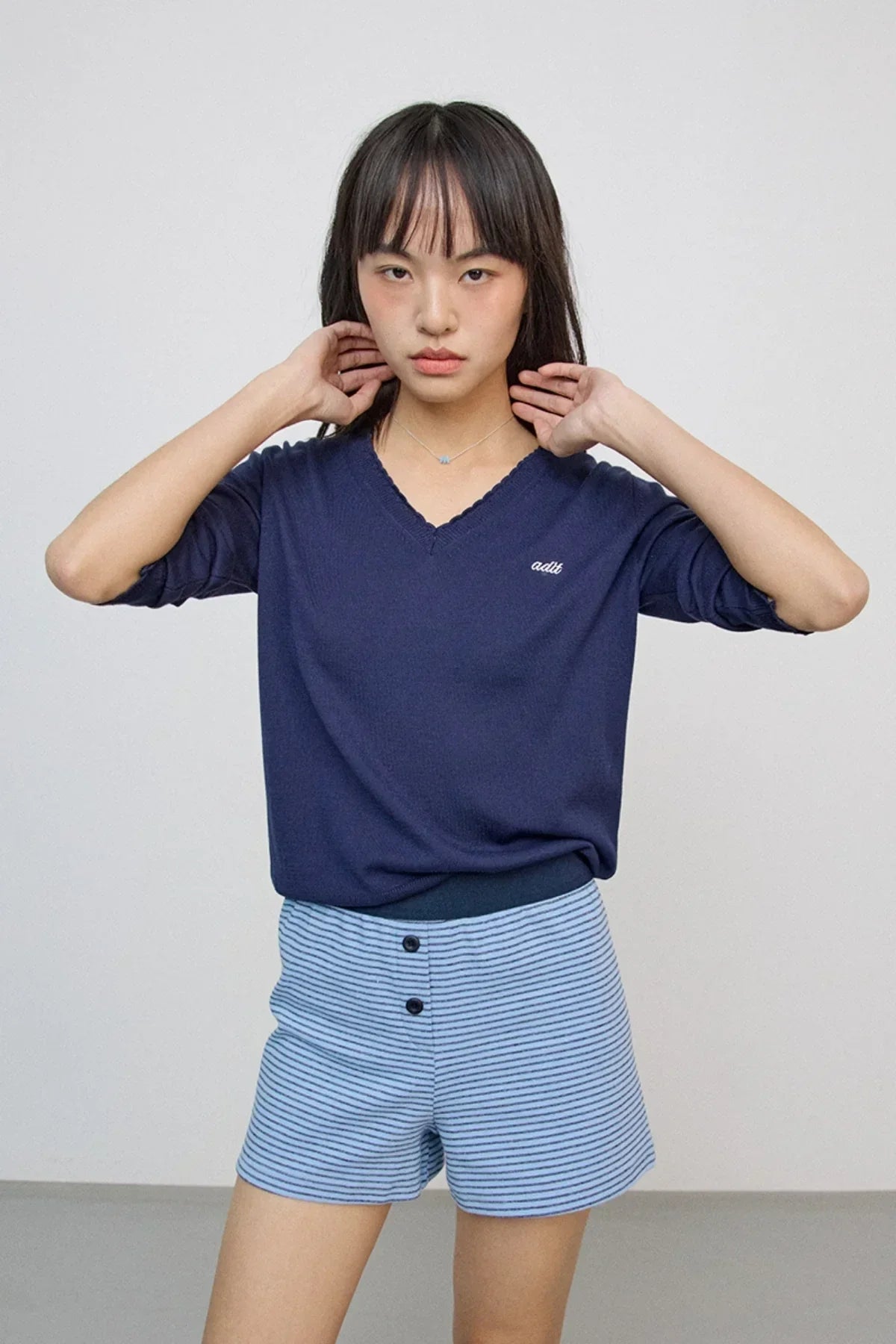 Young woman wearing navy v-neck sweater and light blue striped shorts against plain background