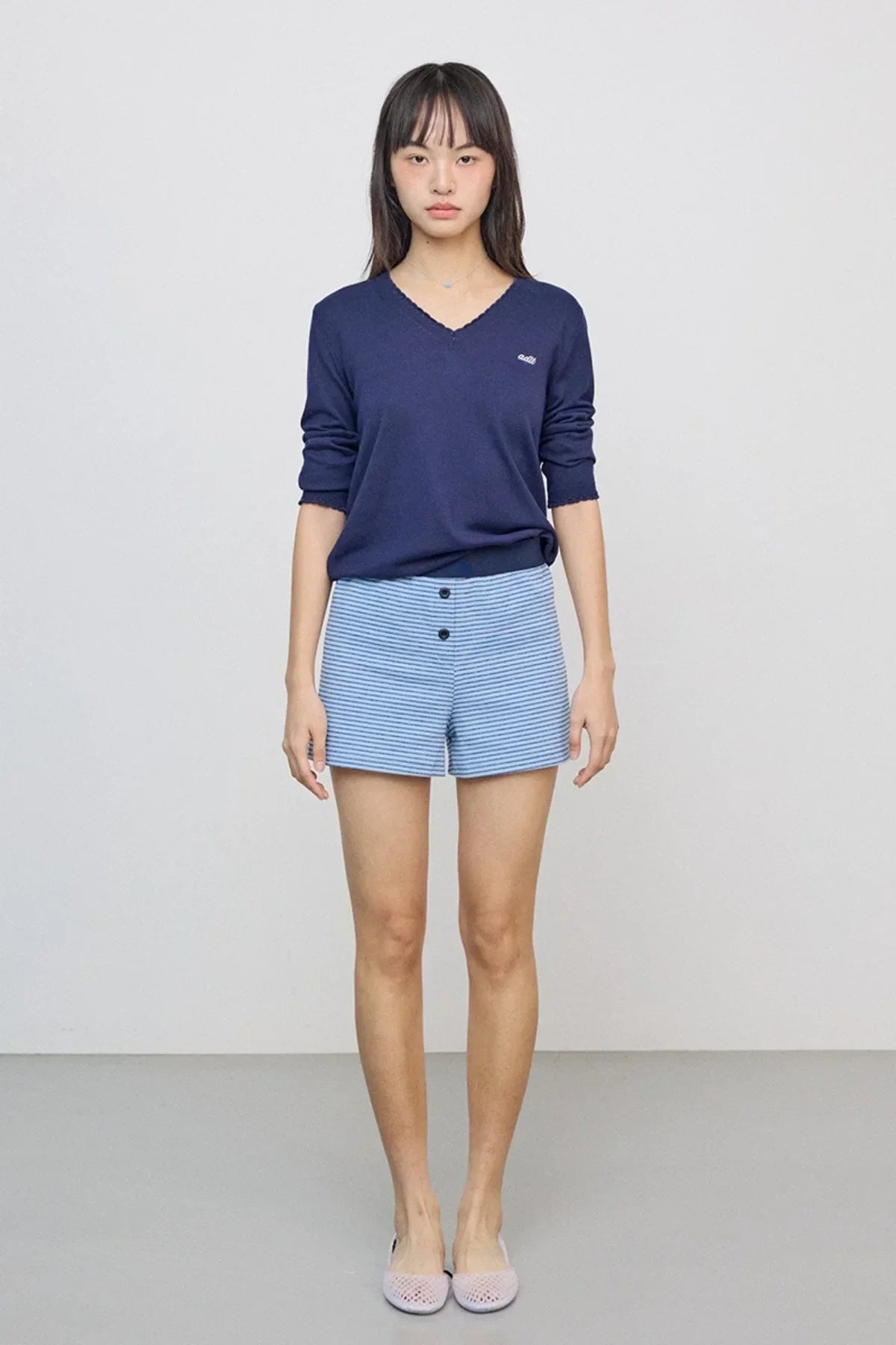 Woman wearing navy V-neck knit sweater and light blue striped shorts standing against plain background