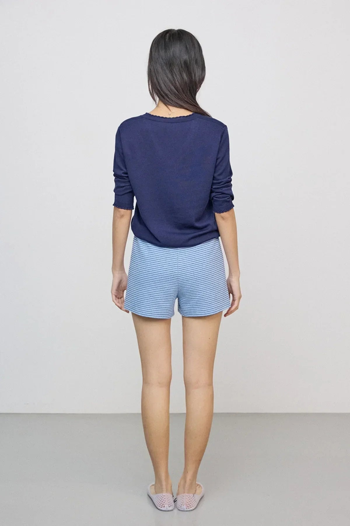 Woman wearing navy long-sleeve top and light blue striped shorts, standing against gray background