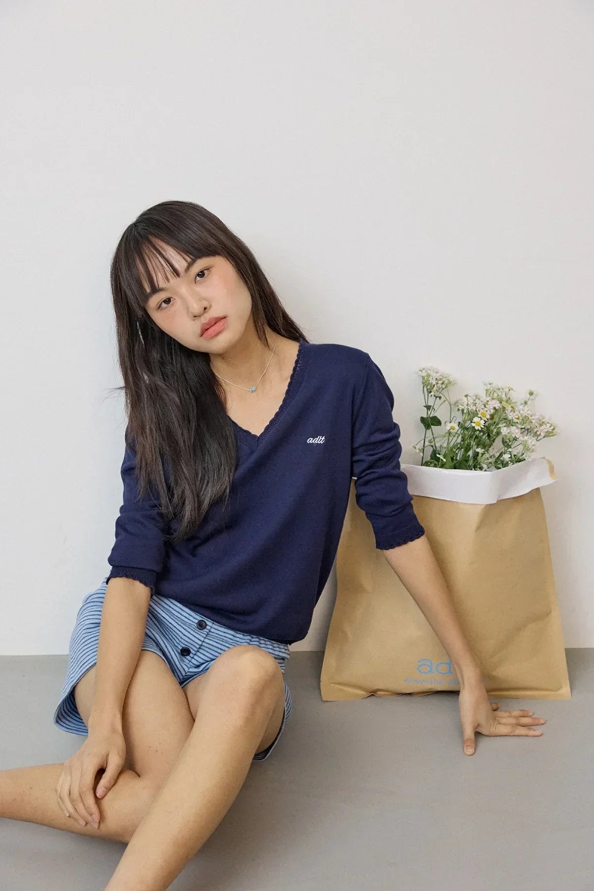 Young woman sitting against white wall wearing navy lace V-neck knit sweater and blue striped skirt