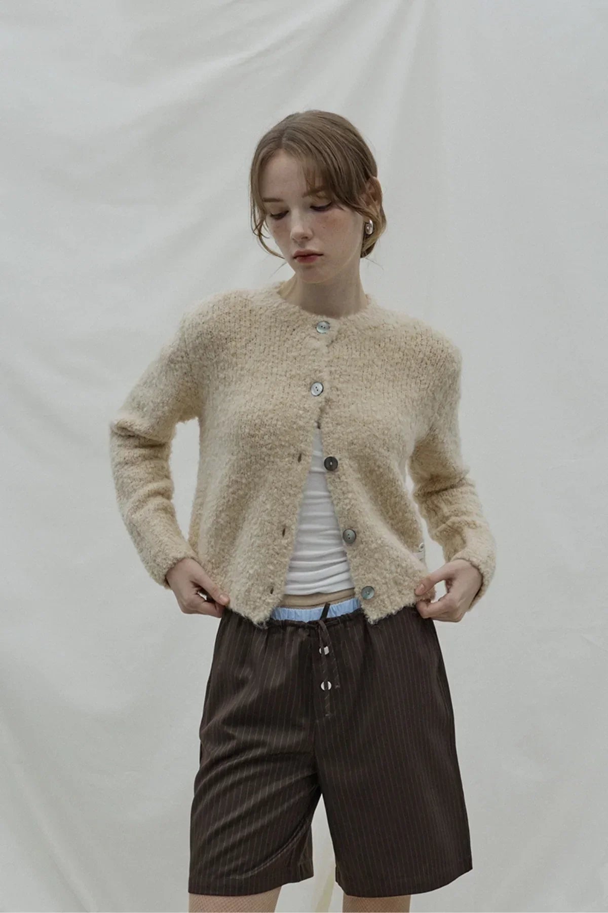Woman wearing beige fuzzy buttoned cardigan and brown pinstripe Bermuda shorts against plain background