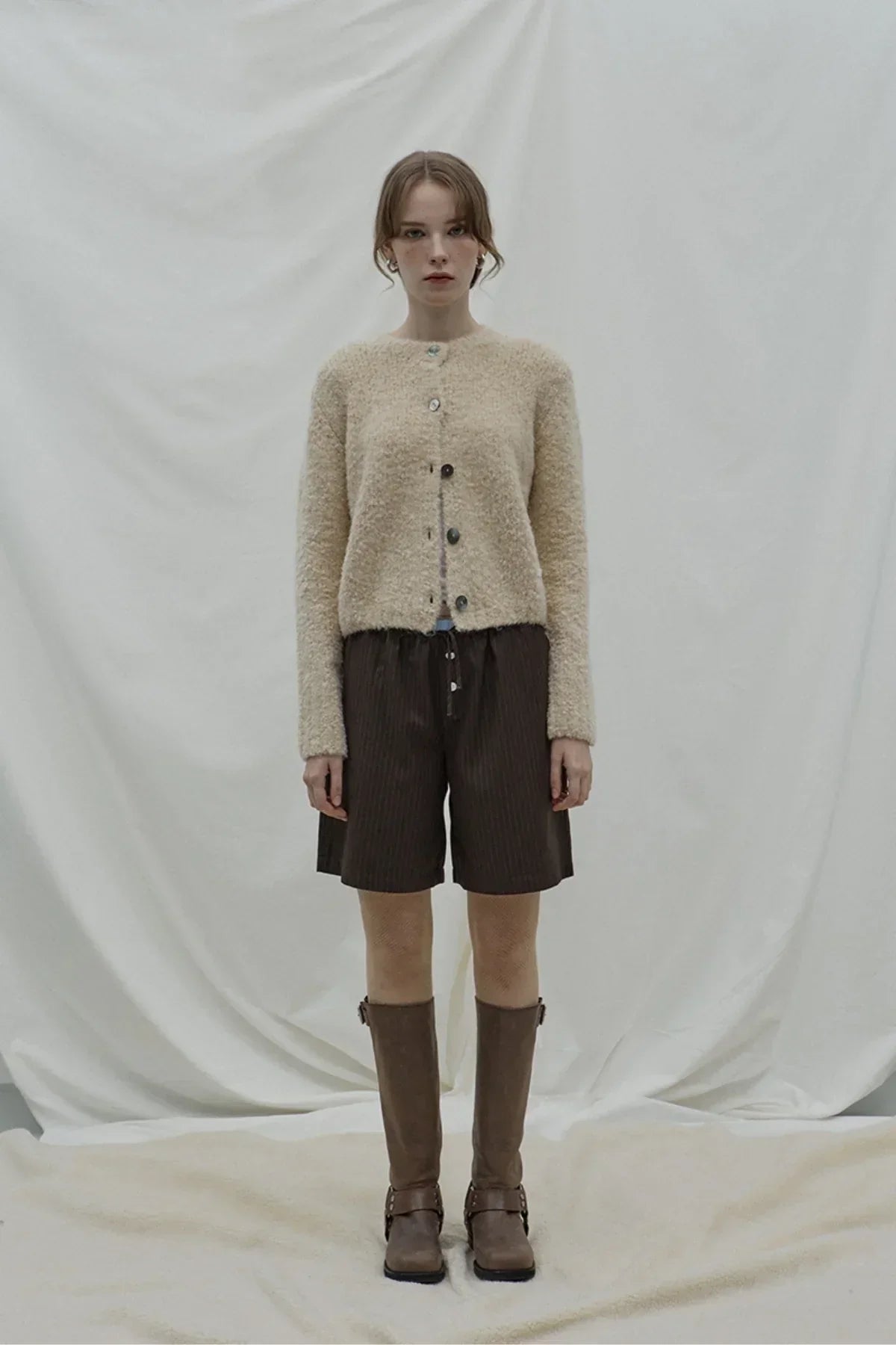 Model wearing beige fuzzy cardigan, brown layered Bermuda shorts, and knee-high brown boots on neutral backdrop
