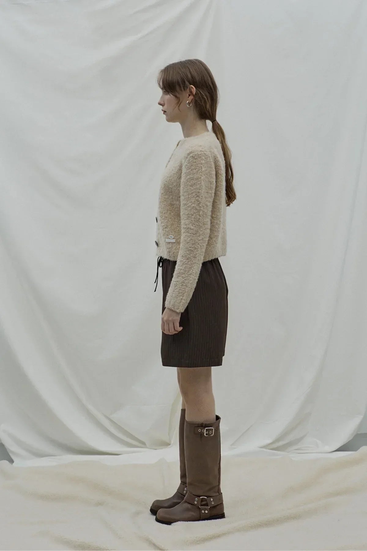 Side view of a woman wearing a beige fuzzy cardigan, dark layered skirt, and tall brown boots against a plain white backdrop