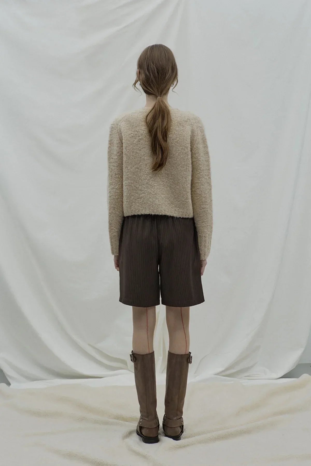 Rear view of woman in beige fuzzy sweater, brown pleated Bermuda shorts, and knee-high brown boots