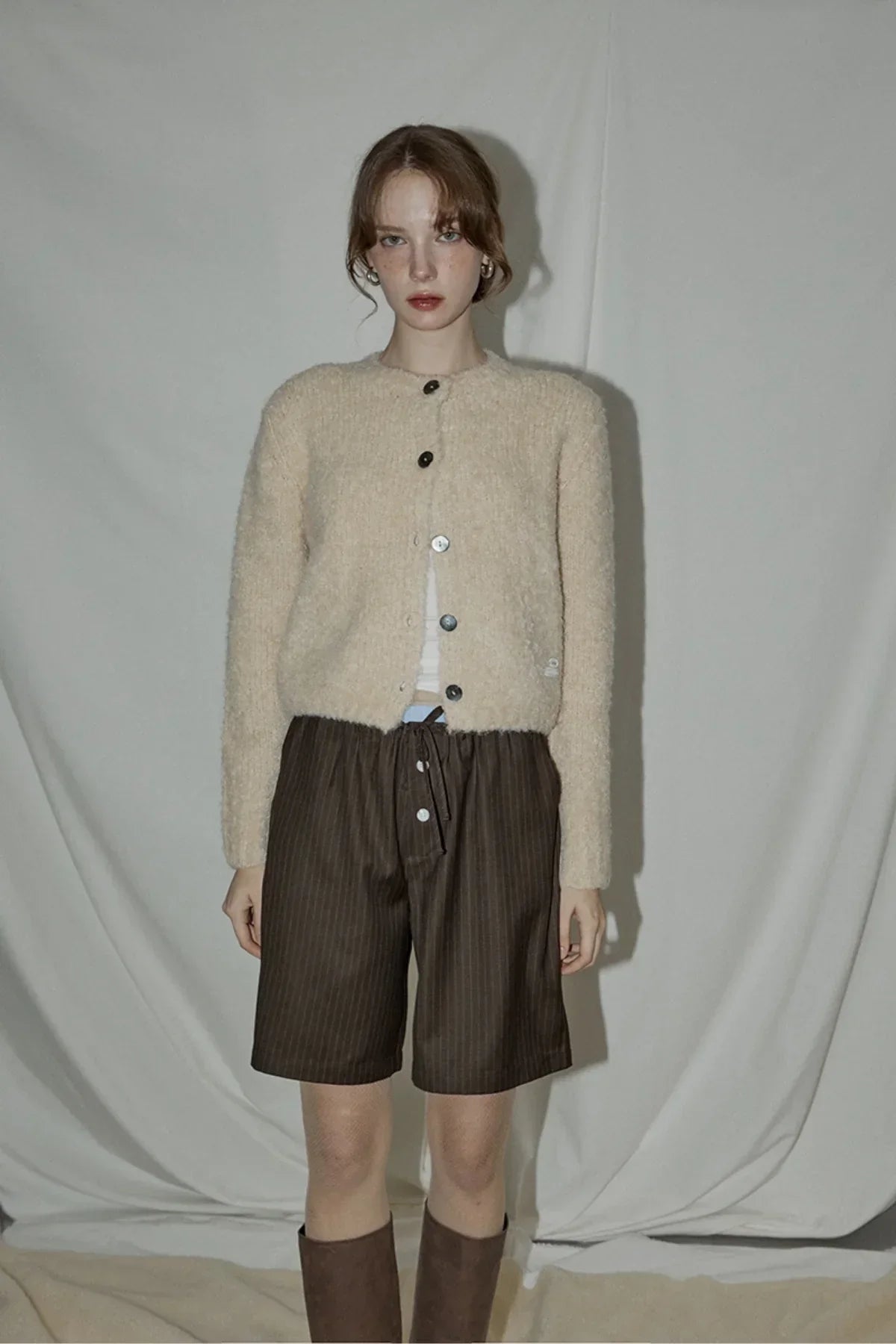 Model wearing beige fuzzy cardigan and brown pinstripe layered Bermuda shorts in studio