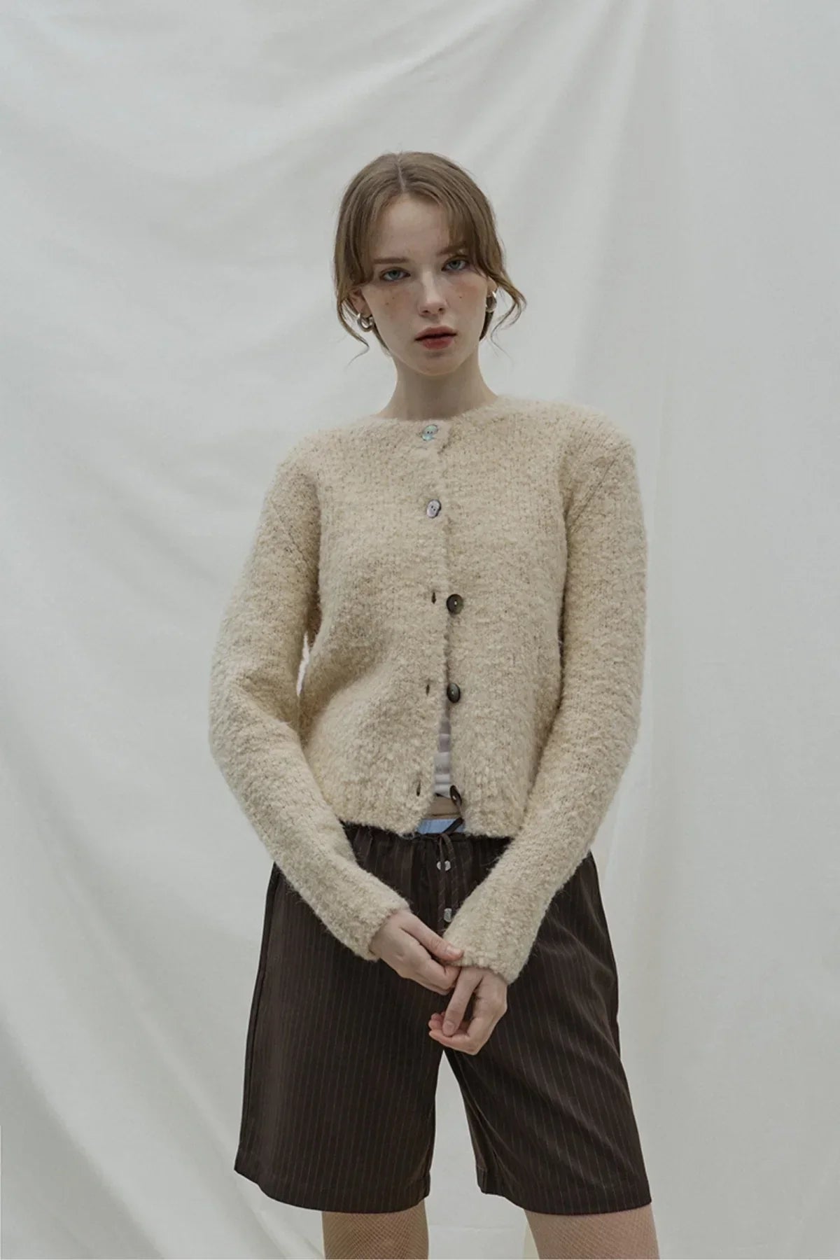 Young woman in beige fuzzy cardigan and brown pinstripe layered Bermuda shorts posing in studio
