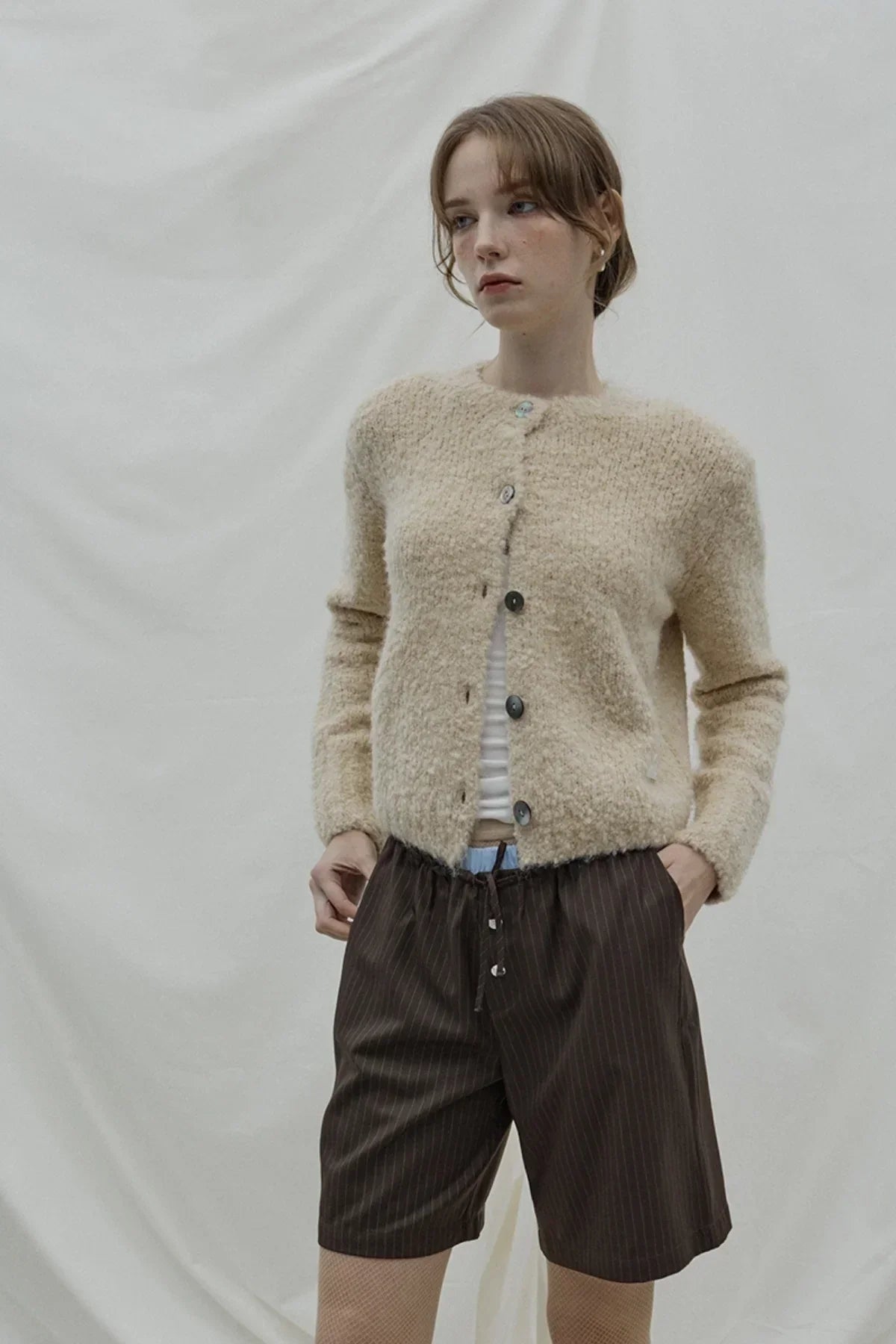 Woman wearing beige fuzzy cardigan and brown pinstripe Bermuda shorts against white fabric backdrop