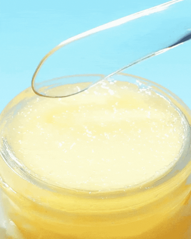 Close-up of yellow lemon sugar scrub lip mask with clear spatula scooping product in jar on blue background