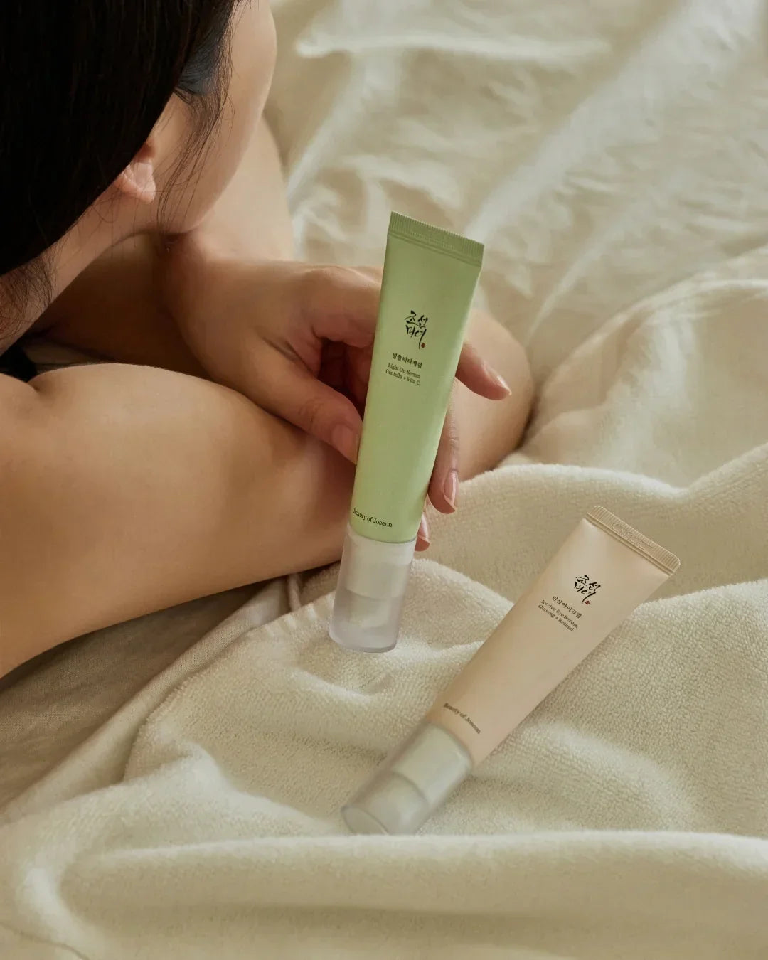 Woman holding green Light On Serum Centella + Vita C skincare tube with another beige tube on beige blanket
