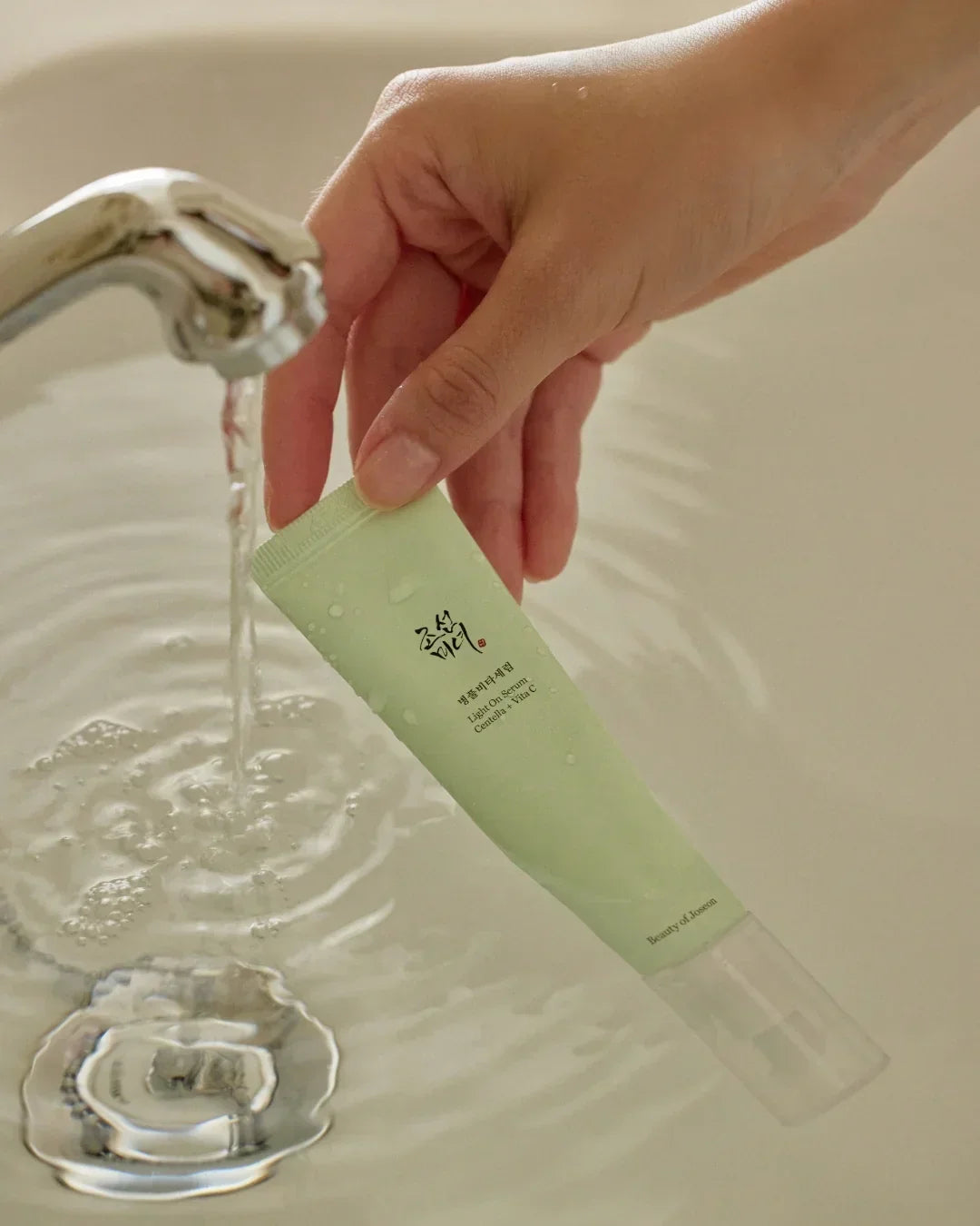 Hand holding green tube of Light On Serum Centella + Vita C under running water in white sink