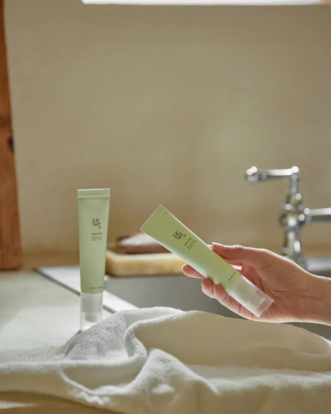 Hand holding green skincare serum tube near sink with second serum and white towel