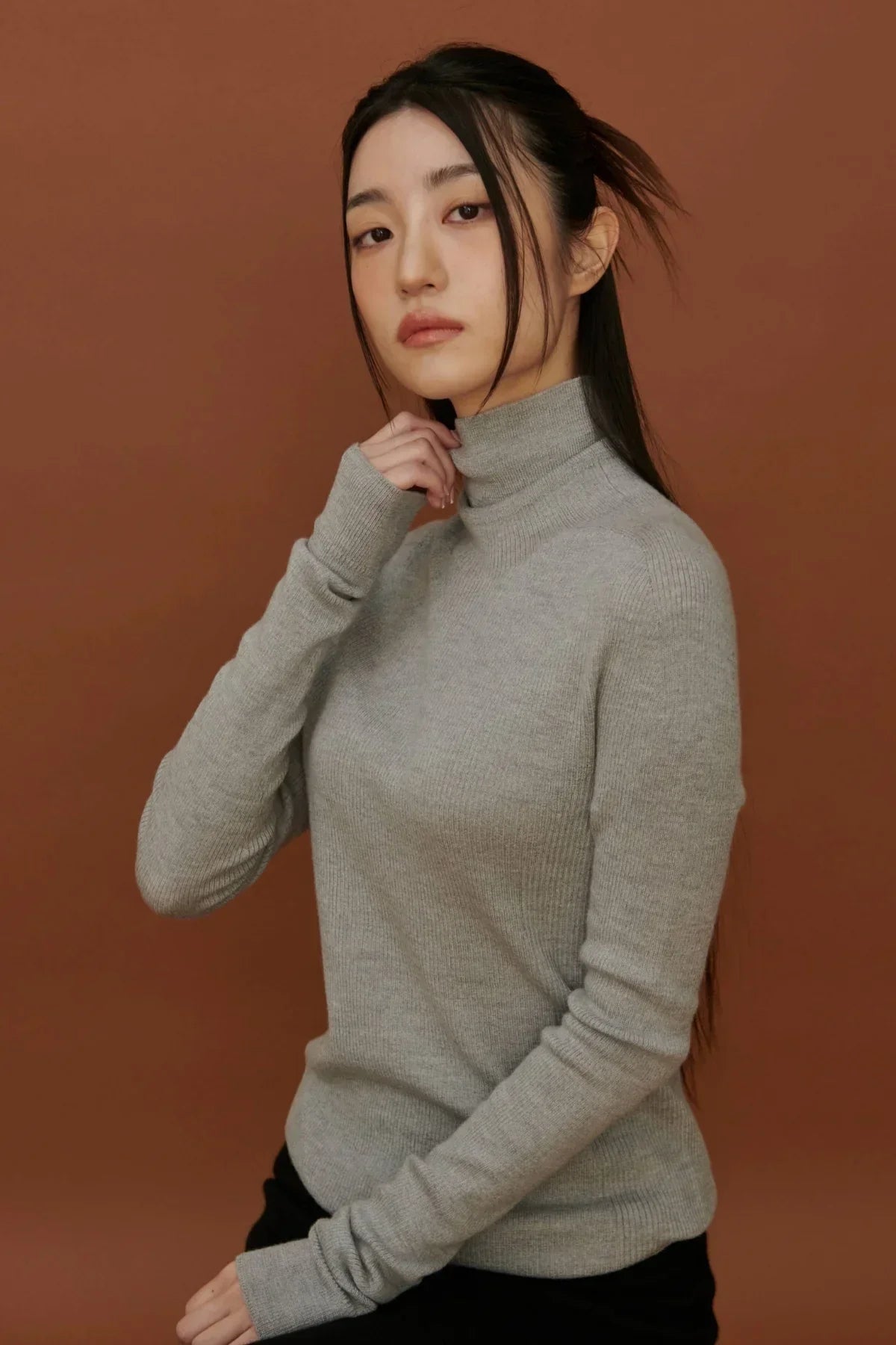 Young woman wearing a light gray lightweight turtleneck knit sweater against a brown background