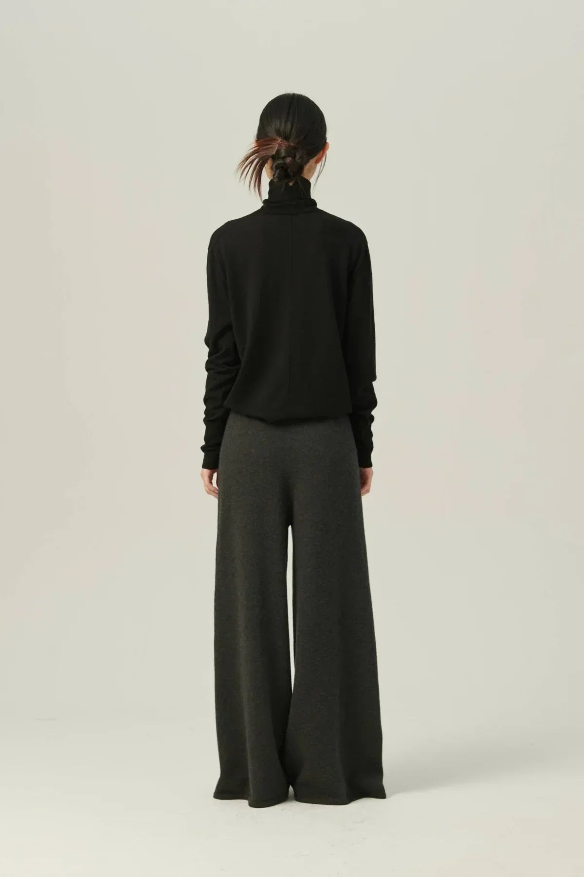 Woman wearing black lightweight turtleneck knit and dark wide-leg pants, standing against plain background