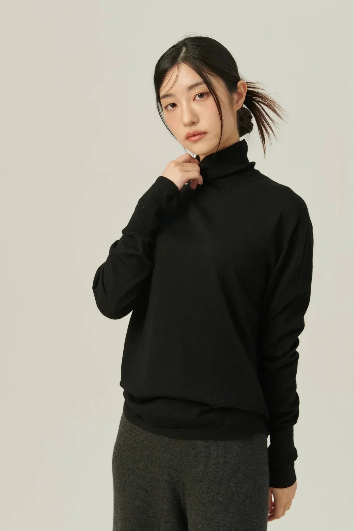 Asian woman wearing a black lightweight turtleneck sweater and dark gray pants against a plain background