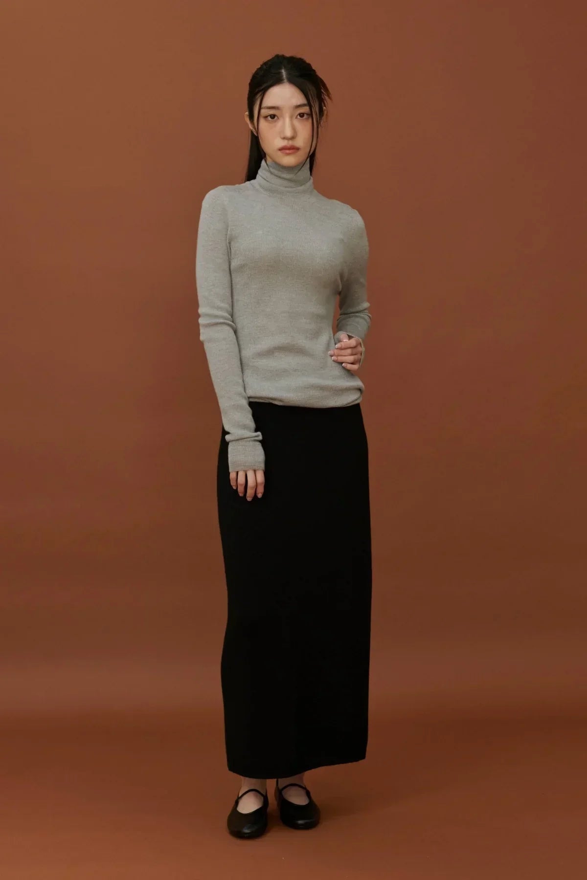 Woman wearing a light grey turtleneck knit sweater and a long black skirt against brown background