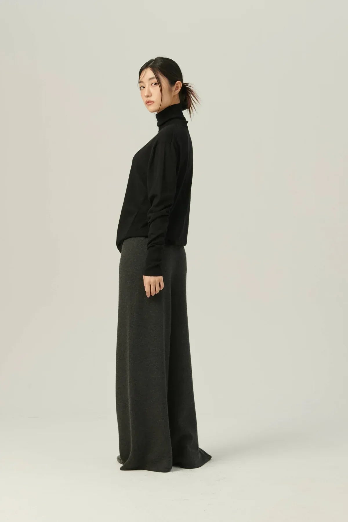 Side view of woman wearing black lightweight turtleneck knit top and wide-leg dark gray pants against light background