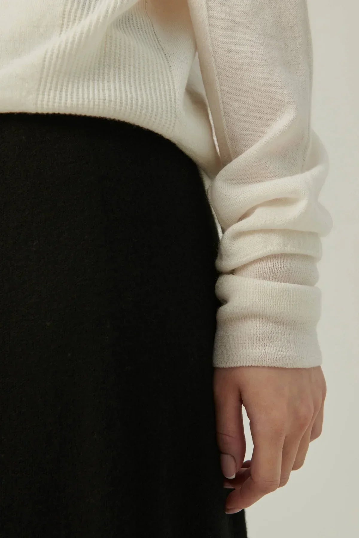 Close-up of a person's hand wearing a lightweight white turtleneck knit sweater with ribbed detailing and black skirt