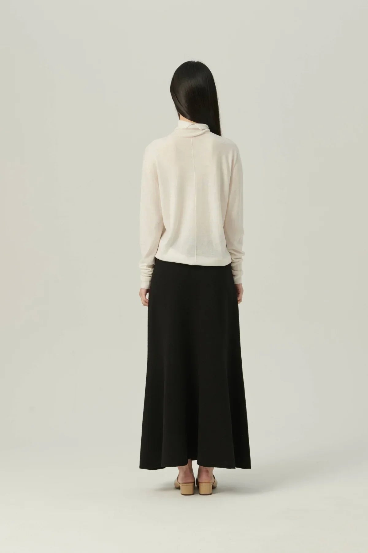 Woman wearing lightweight white turtleneck knit sweater and long black skirt standing against plain background