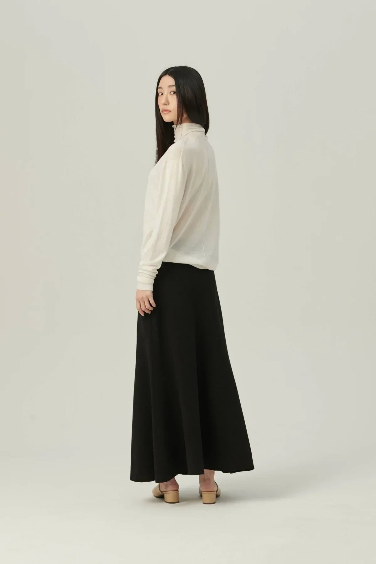 Woman in white lightweight turtleneck knit sweater and black maxi skirt against plain background