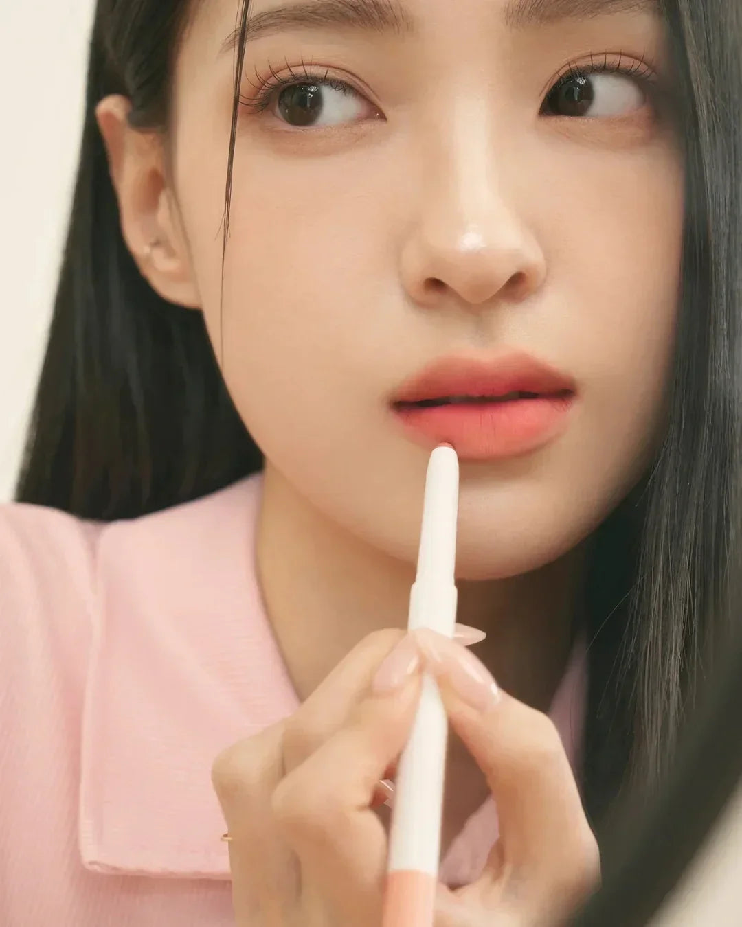 Close-up of a woman applying coral lip pencil with long black hair and pink shirt